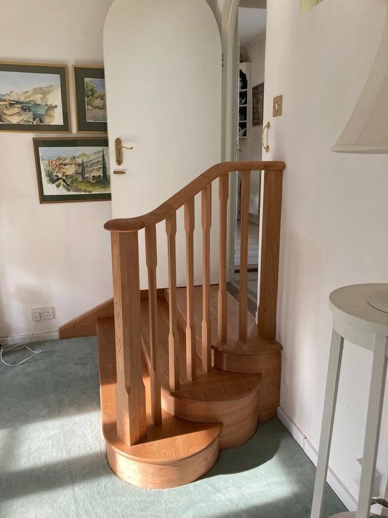 Wooden spiral staircase in a room with white walls, green carpet, and framed landscape paintings.