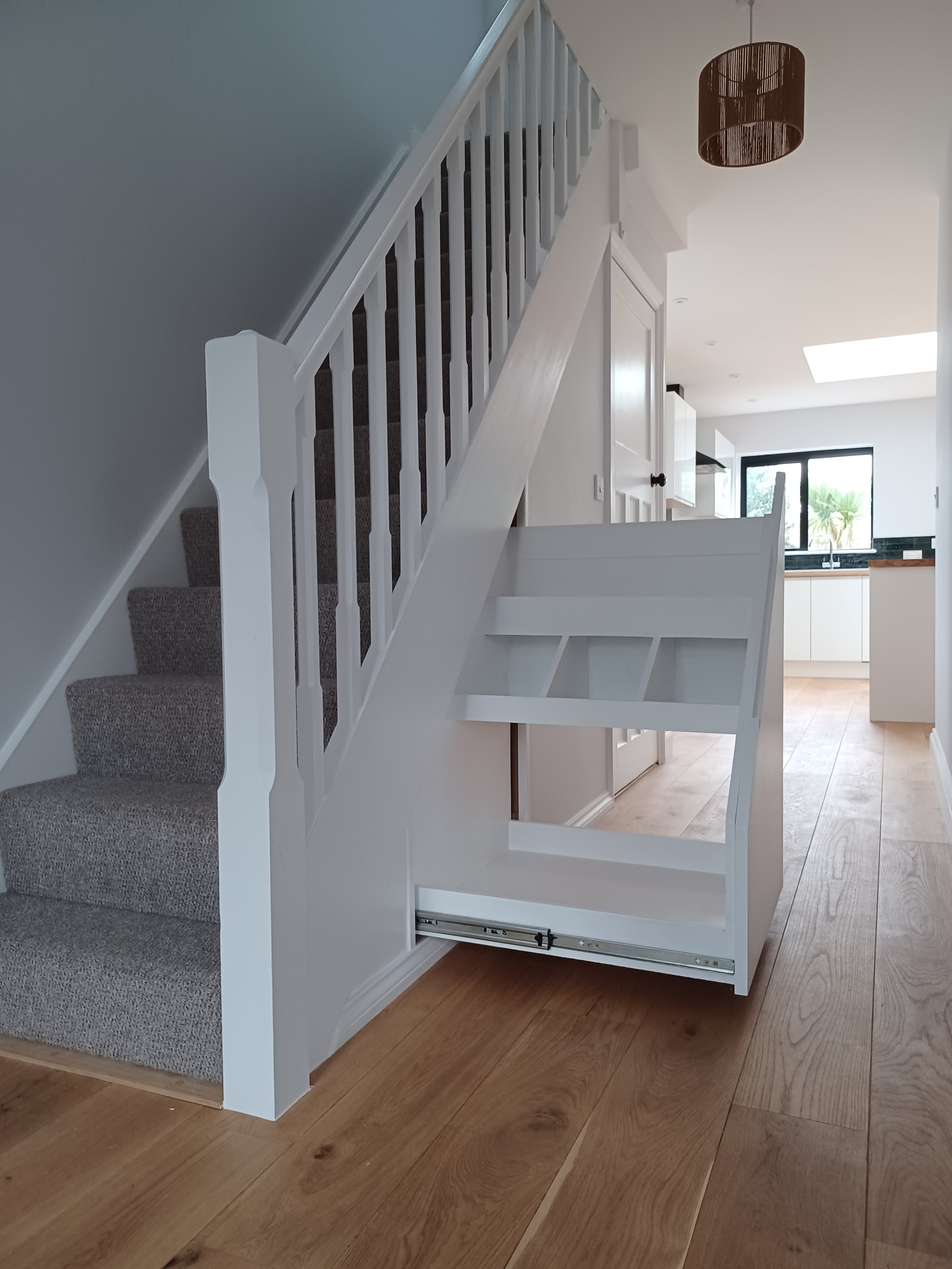 A staircase with a carpeted step leading up, and the lower part is under construction with a white panel and partial frame. The background shows a bright kitchen with light wood flooring, white cabinets, and a large window.