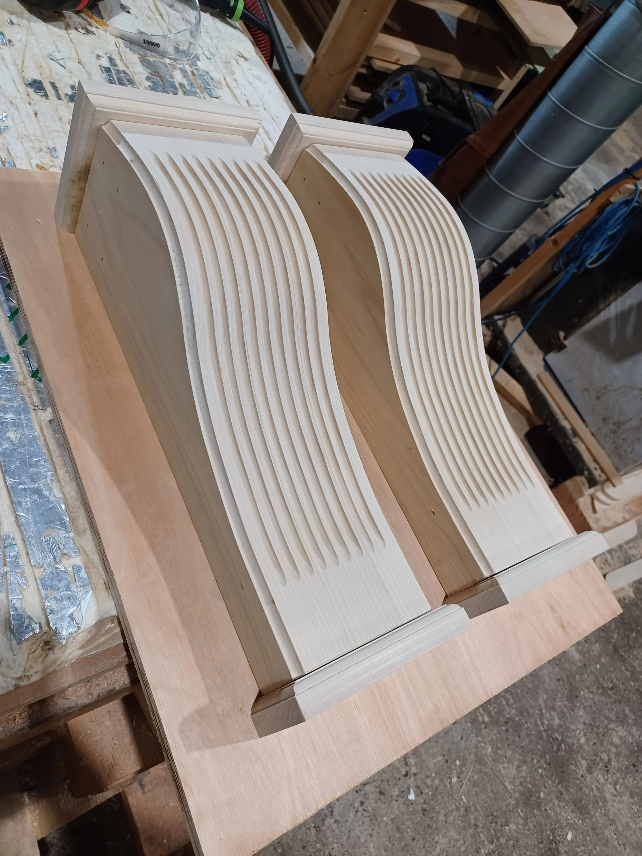 Two unfinished wooden speaker cabinets with curved, slotted fronts on a workbench in a woodworking shop.