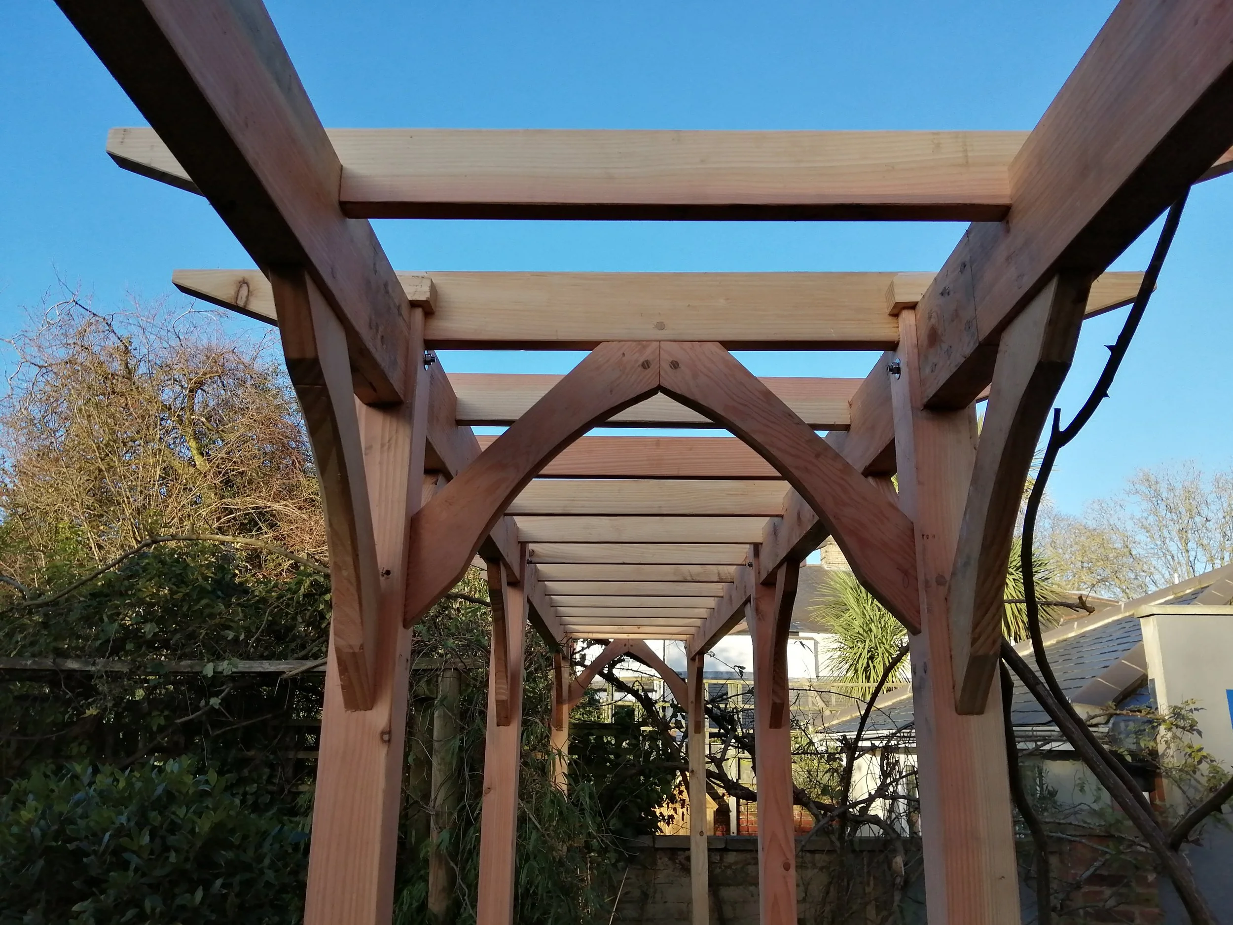 A wooden pergola under construction with beams and supports in a backyard with trees and sky.