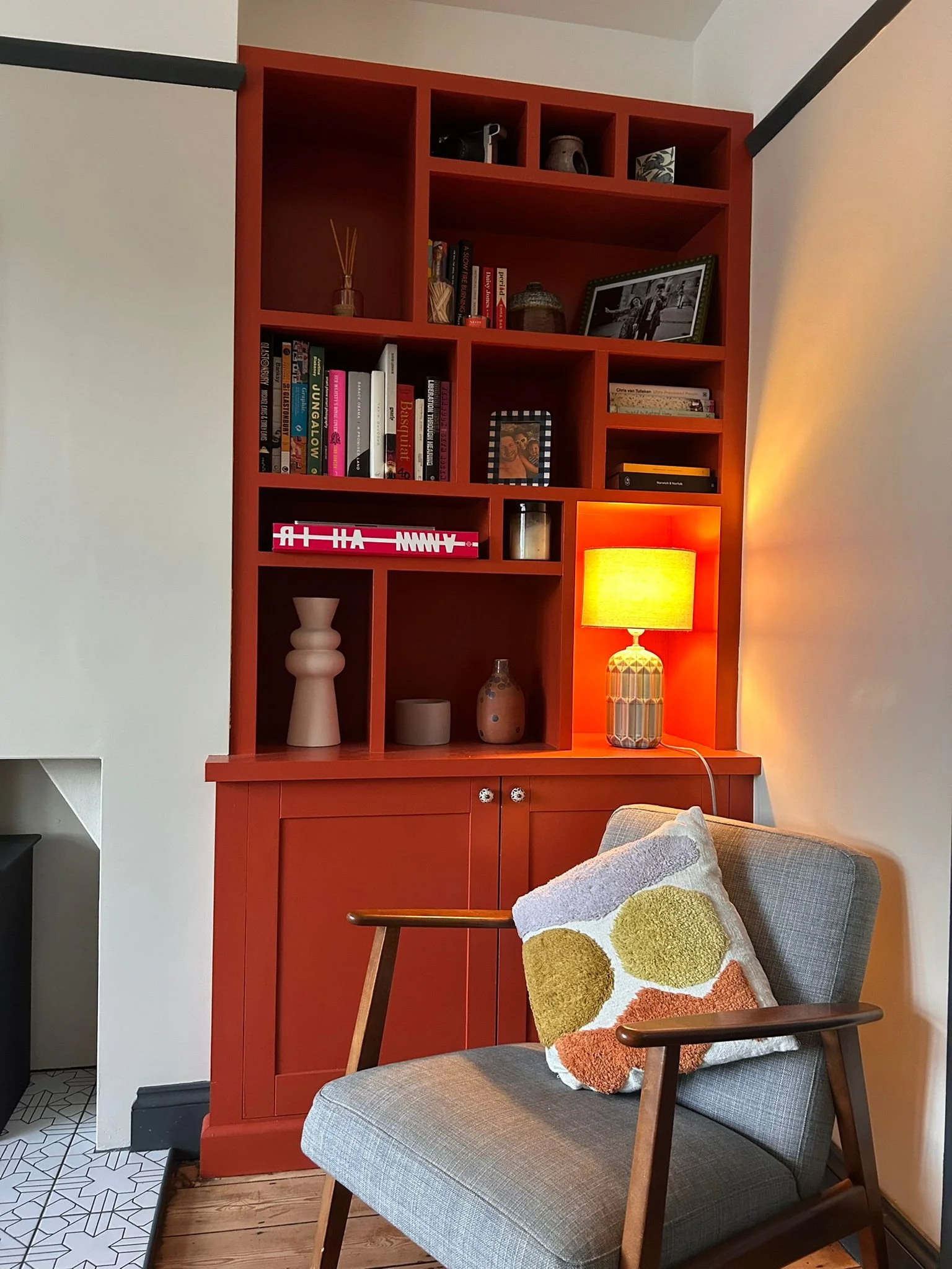 A cozy living room corner with a gray armchair that has wooden armrests and a decorative multicolored cushion. Behind the chair is a built-in red bookshelf with various books, decorative objects, a framed photograph, and a table lamp emitting warm li