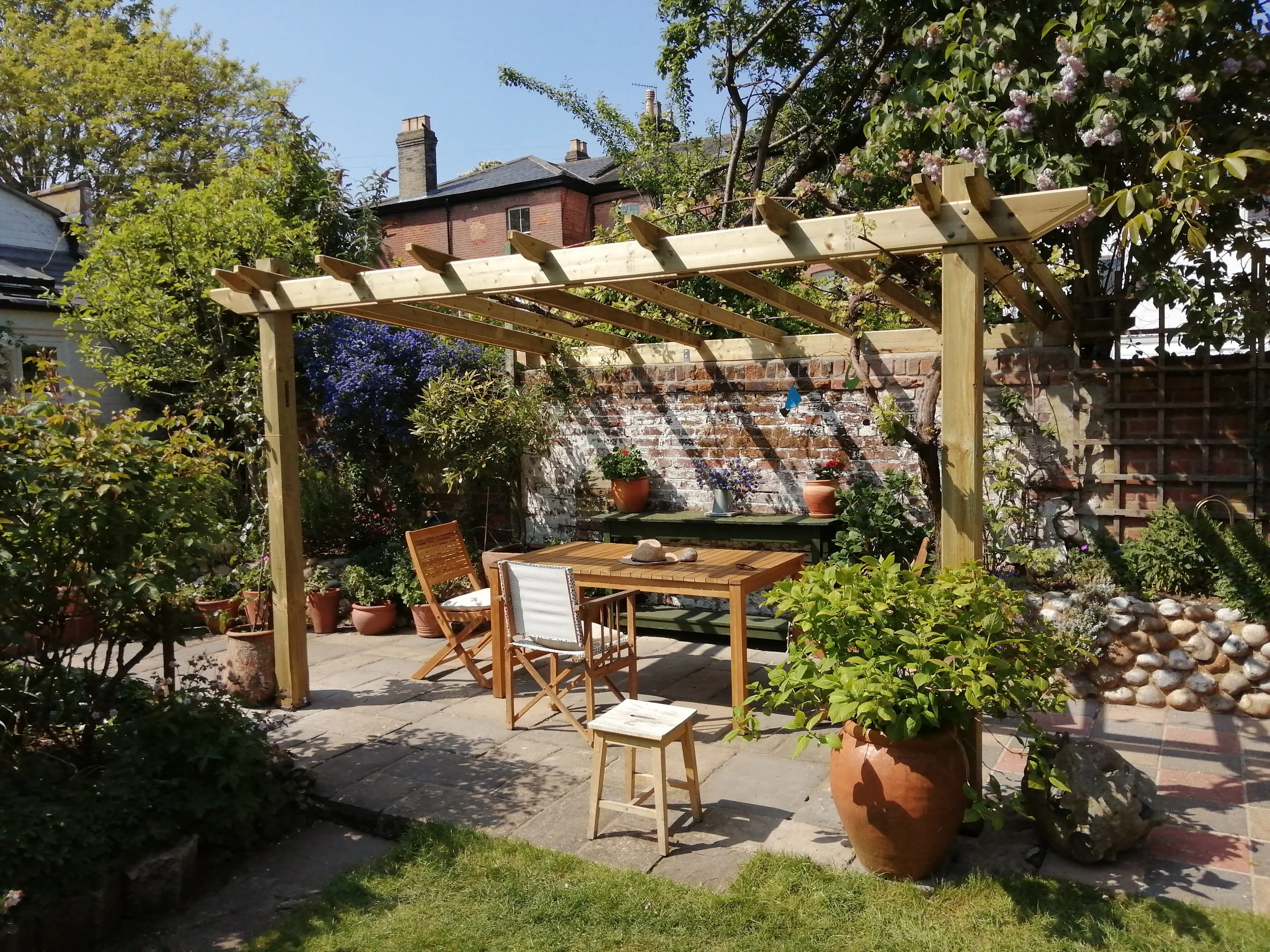 A lush backyard patio with potted plants and a wooden pergola. There is a dining table with chairs, surrounded by greenery and flowering bushes, with a brick wall in the background and a clear blue sky overhead.
