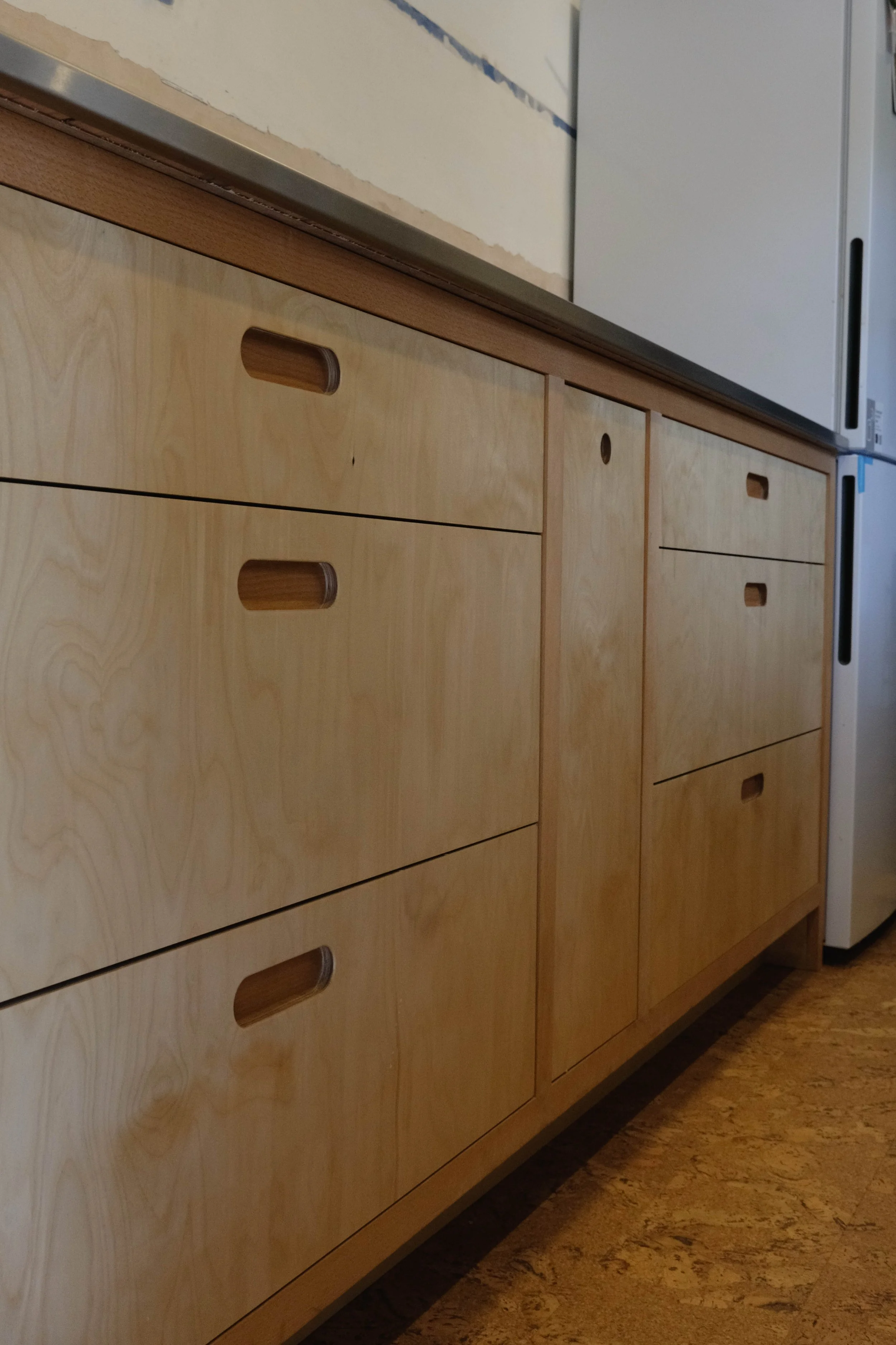Wooden kitchen cabinet drawers with integrated handles, next to a white refrigerator and a counter surface.