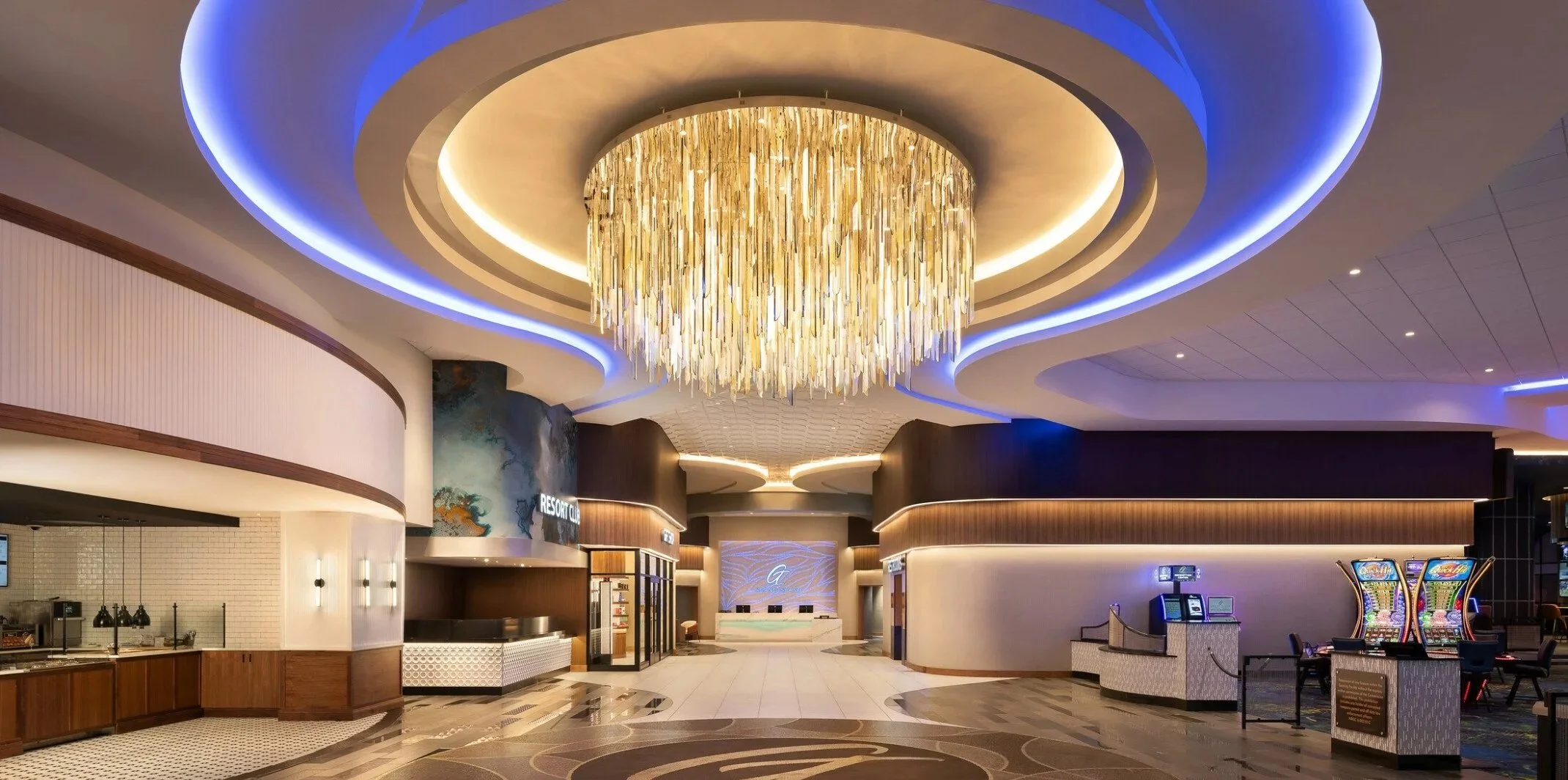 Interior of a modern resort casino lobby featuring a large decorative crystal chandelier, illuminated ceiling design with blue and warm lighting, a check-in desk with a bright digital sign, slot machines, and a neutral color palette with warm wood accents.