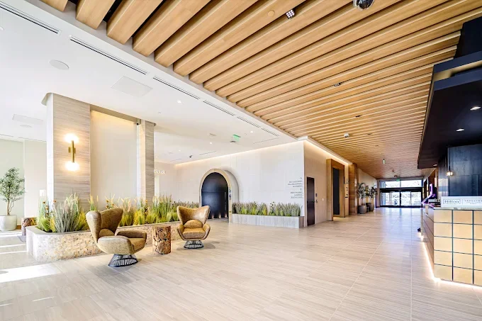 Modern hotel lobby with wooden ceiling beams, beige chairs, and green plants.