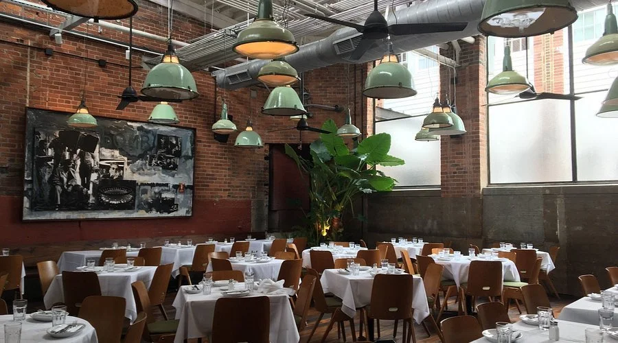 Elegant restaurant with round tables covered in white tablecloths, set with glasses and silverware, surrounded by wooden chairs, decorated with green hanging pendant lights and a large plant, with exposed brick walls and large windows.