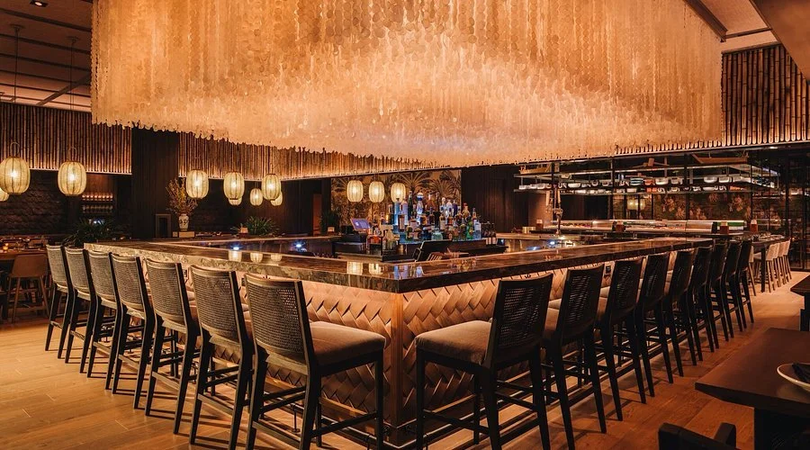 Empty upscale bar with a square-shaped counter, bar stools, hanging pendant lights, and a large decorative chandelier overhead.