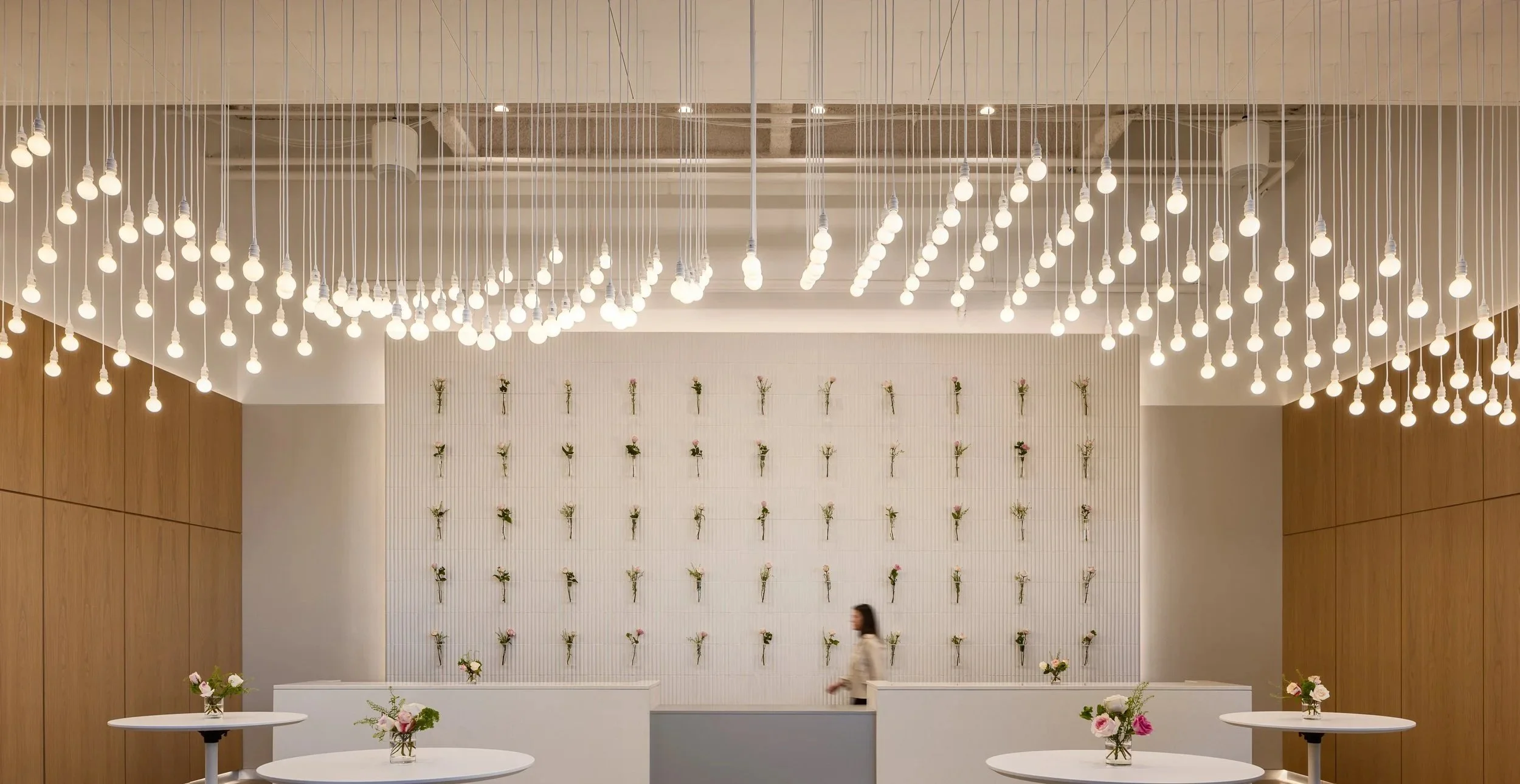 Modern floral display with hanging light bulbs and white tables with flower bouquets in a spacious, well-lit room.