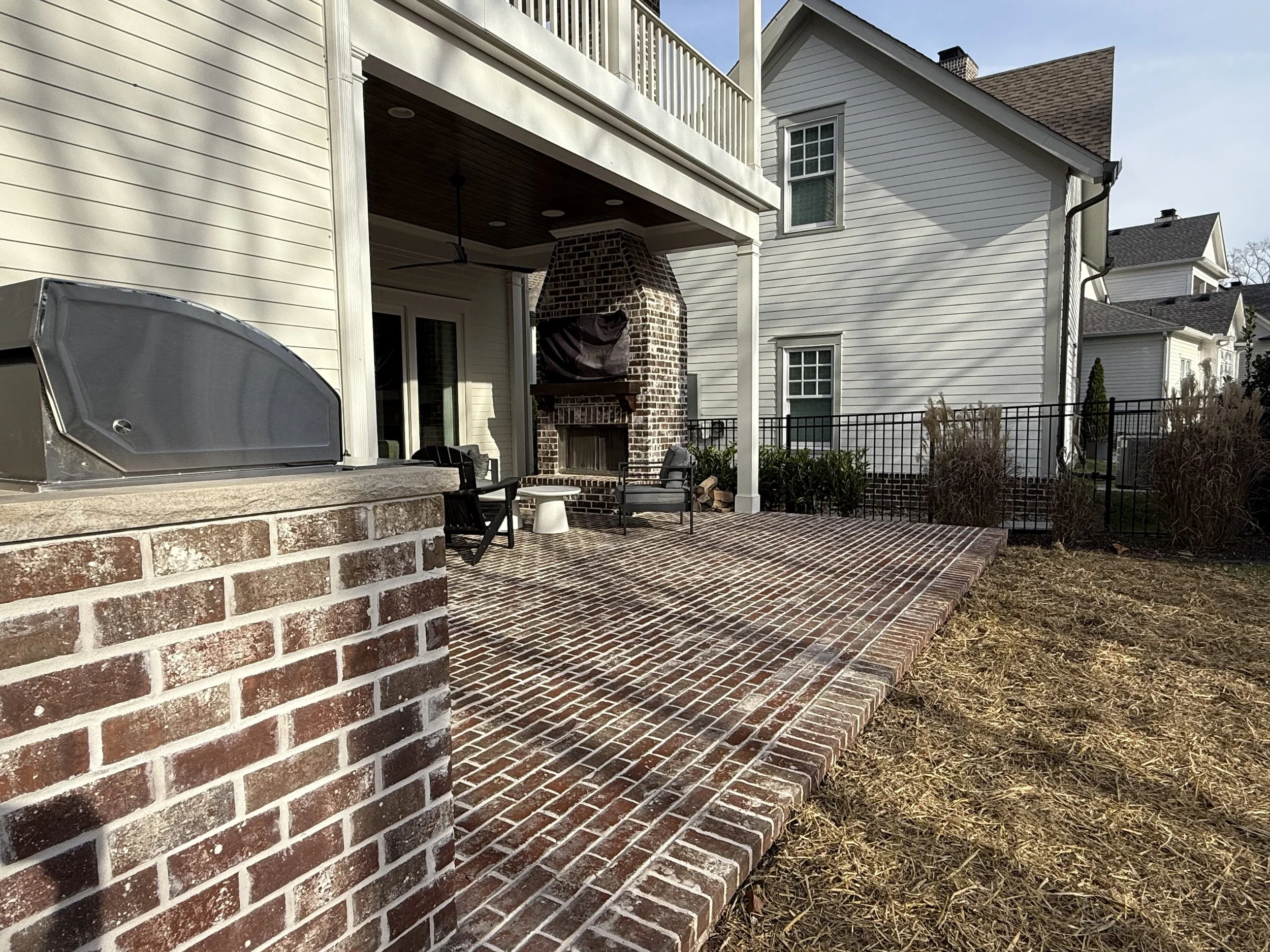 Backyard patio area with brick flooring and brick fireplace, two black chairs with cushions, a small table, a grill, and a porch ceiling fan under a covered porch, surrounded by neighboring houses.