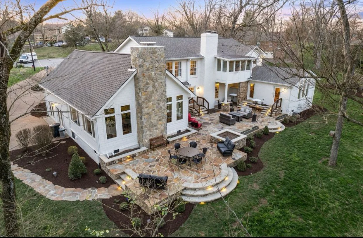 A large white house with multiple levels, stone chimneys, and a spacious outdoor patio area with various seating arrangements, viewed from above during the evening.