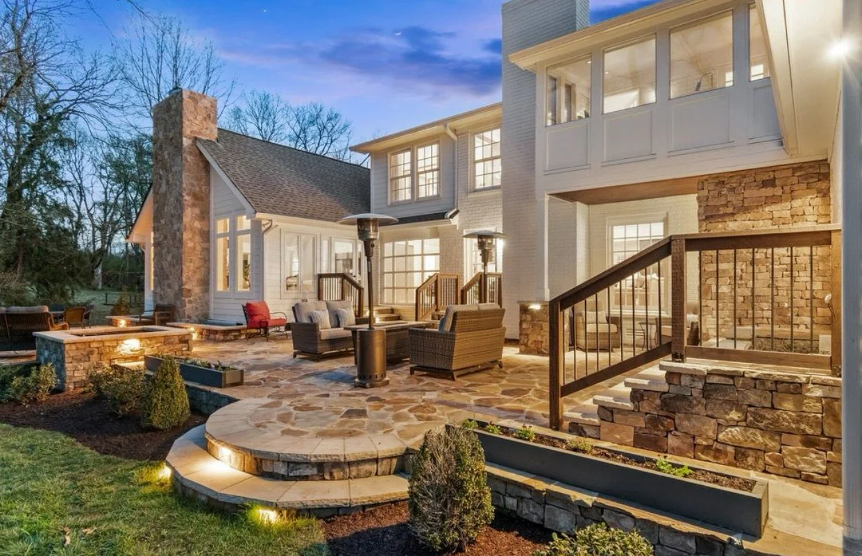 Exterior view of a spacious multi-story house with a stone and white siding facade, large windows, and an illuminated backyard patio with outdoor furniture and heaters during dusk.