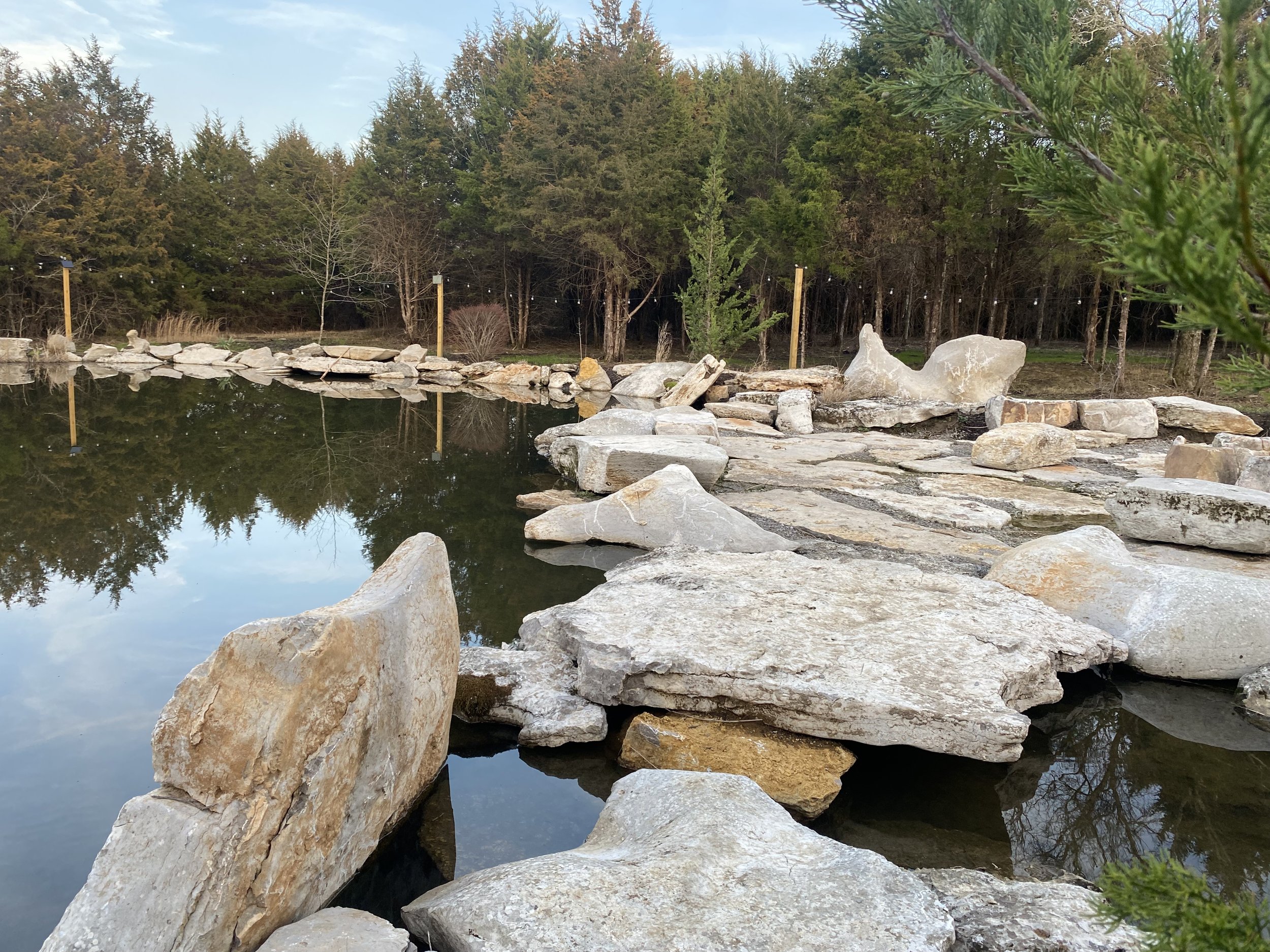 A pond with large rocks along the edge reflecting trees and blue sky, surrounded by greenery and trees in the background.