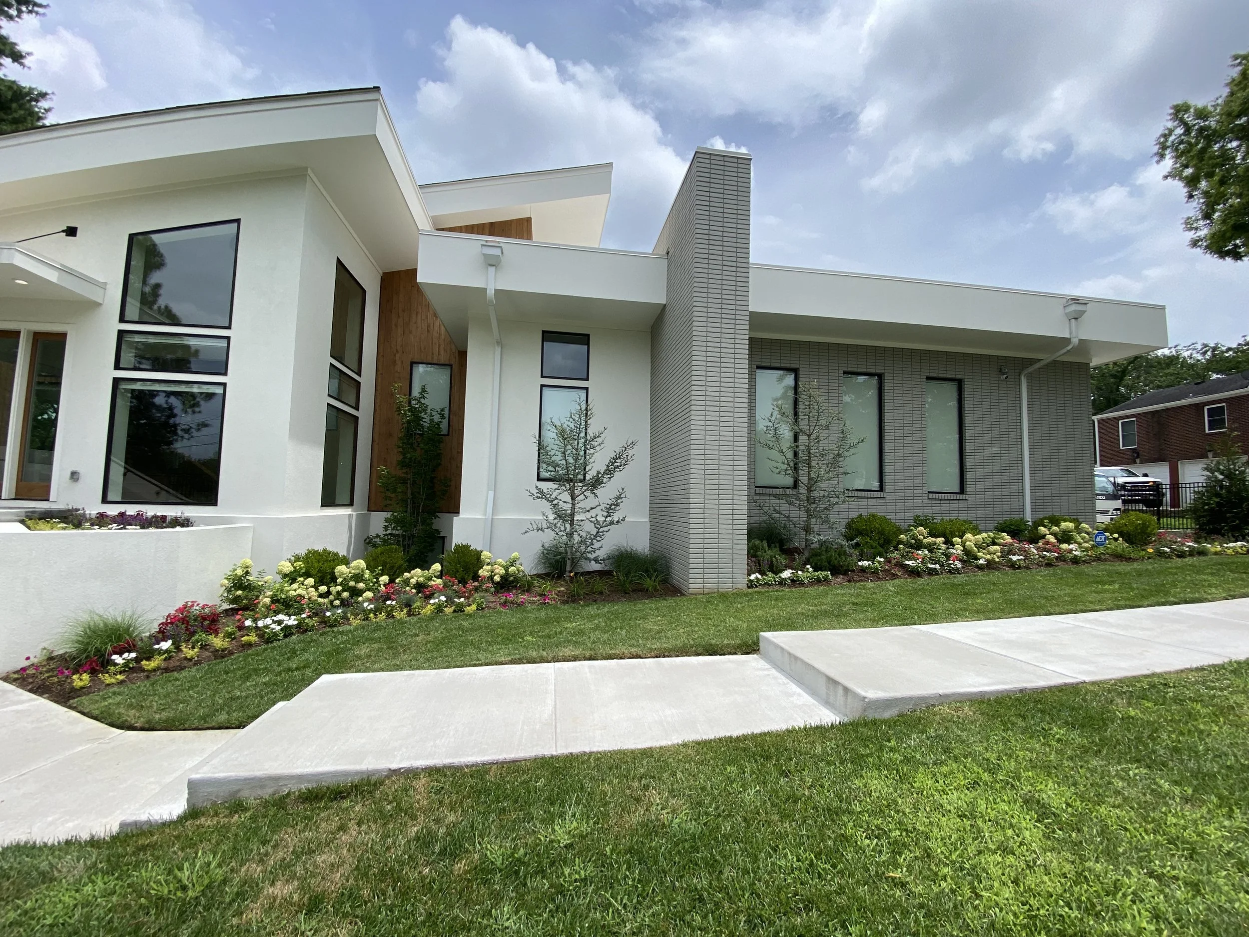 Modern house with white and gray exterior, large windows, landscaped garden with flowers and small trees, concrete sidewalk in foreground, cloudy sky.