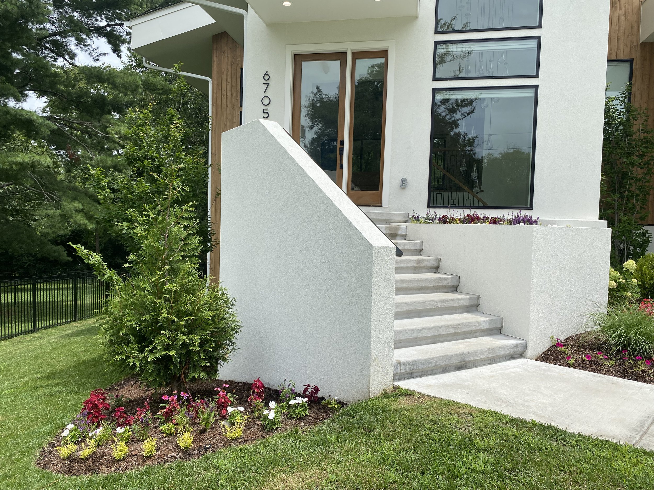 Front view of a modern house with white exterior, brown front door, and large glass windows. Concrete stairs lead up to the entrance, flanked by landscaped flower beds with small plants and bushes. The house number "9705" is visible beside the door.