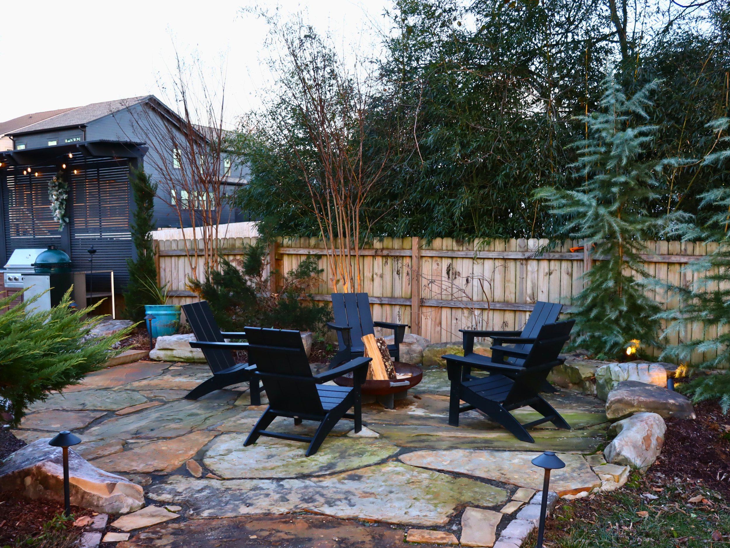 Backyard patio with four black Adirondack chairs arranged in a circle around a fire pit, surrounded by large rocks and lit by small ground lights, with a wooden fence, trees, and a house in the background.