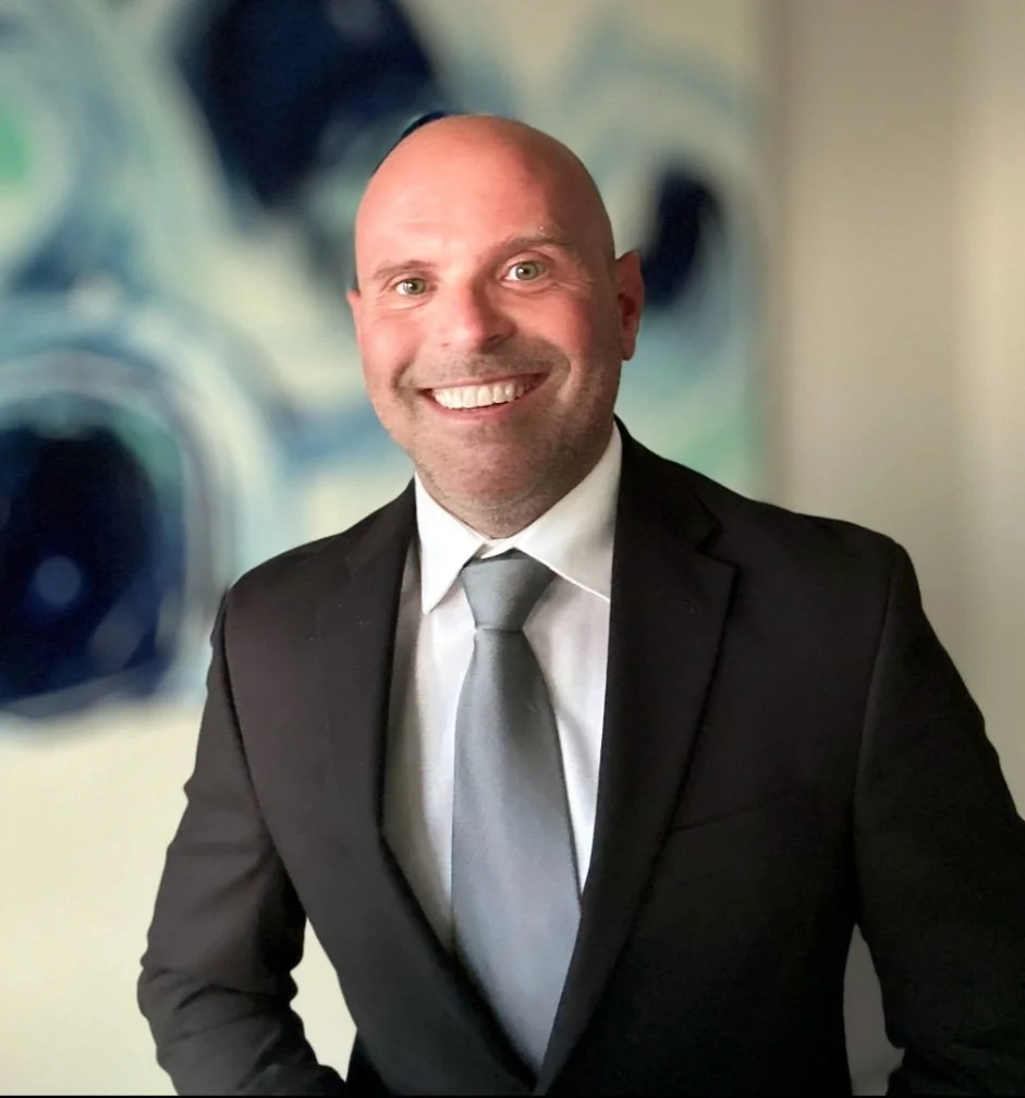 A smiling man with a bald head wearing a black suit, white shirt, and gray tie stands in front of a blurred background that resembles washing machines.