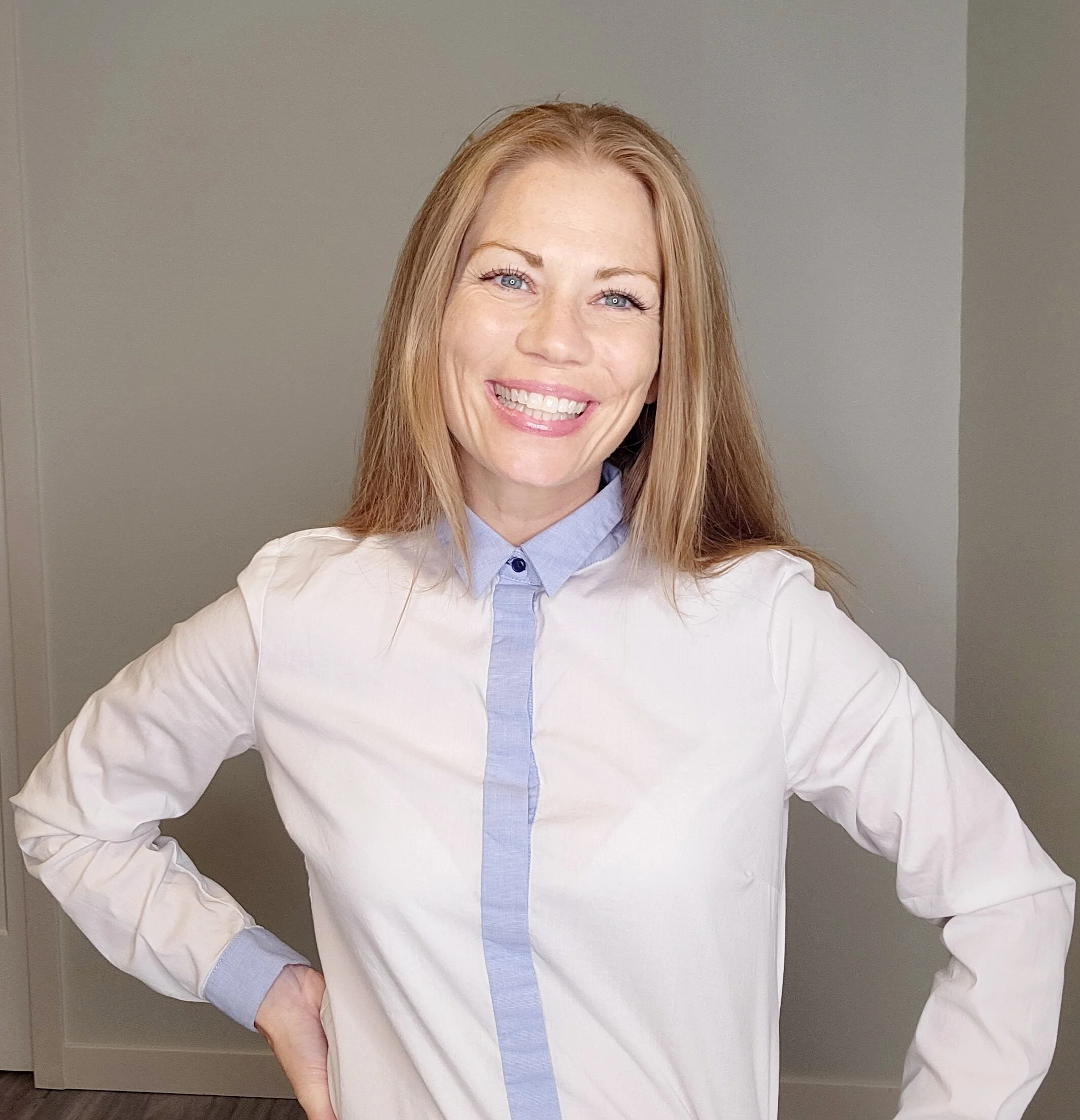 A woman with red hair smiling, wearing a white shirt with light blue details, standing indoors.