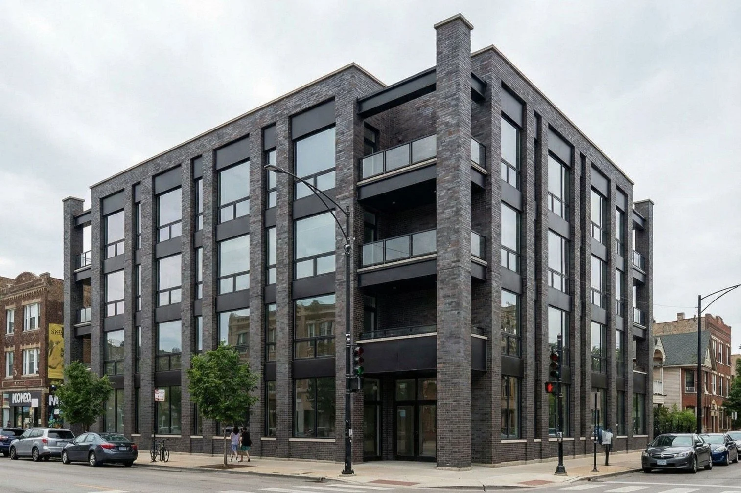 Modern 4-story apartment building with dark brick exterior, large windows, and small balconies at each level, situated on a city street with trees and cars.