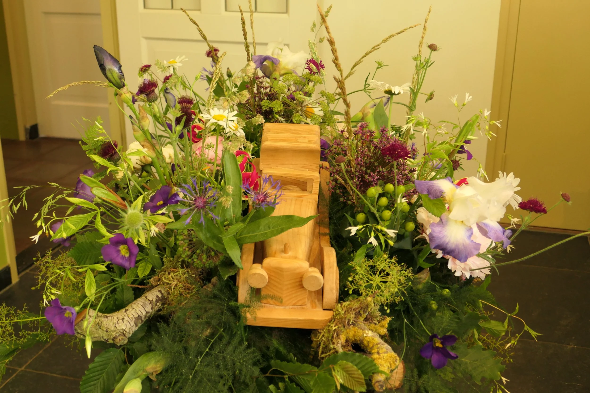 Een bloeiende bloemenarrangement met diverse kleurrijke bloemen en groene bladeren, inclusief een klein houten treinmodel in het midden.