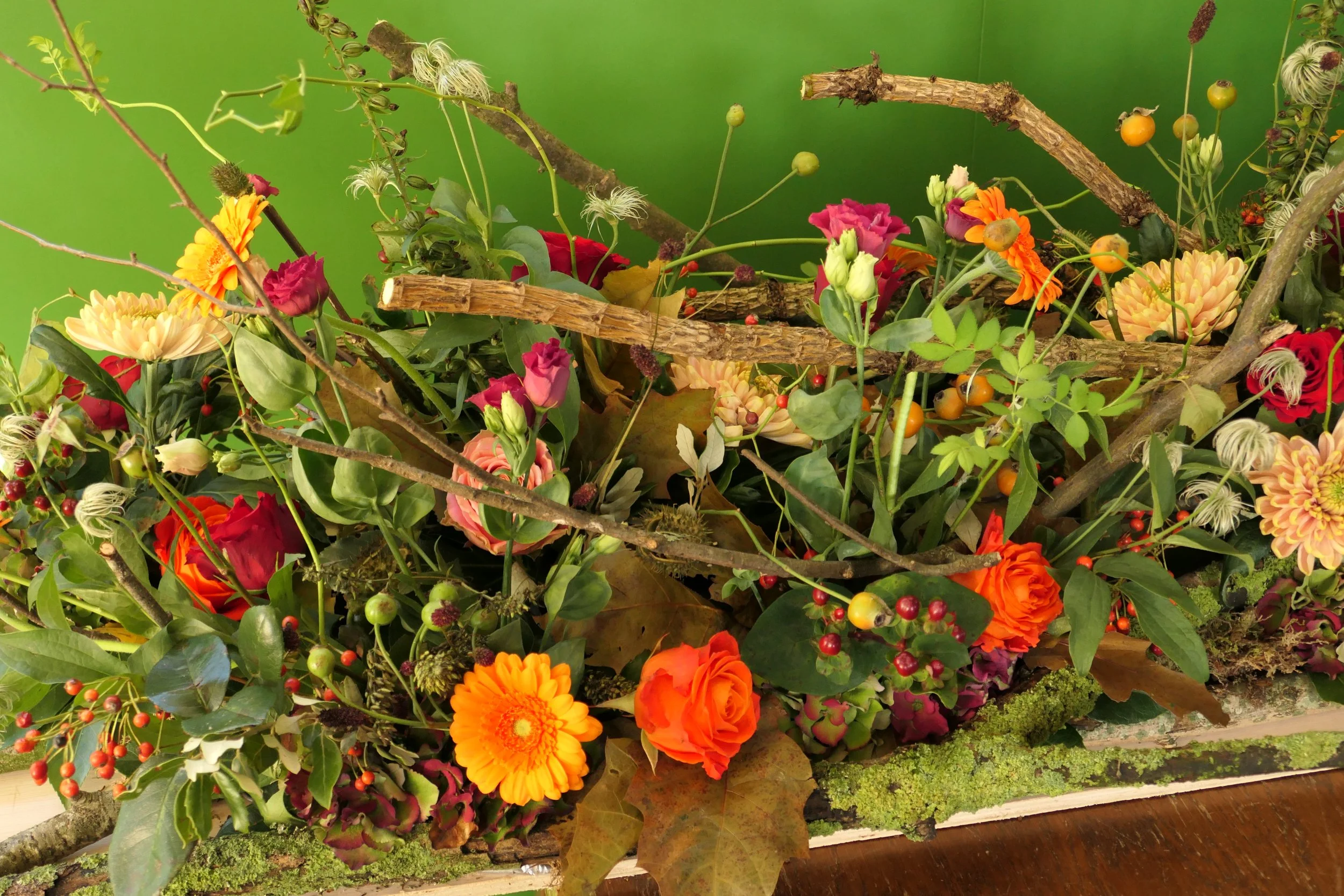 Bloemstuk met diverse bloemen, takken en groene bladeren tegen een groene achtergrond.