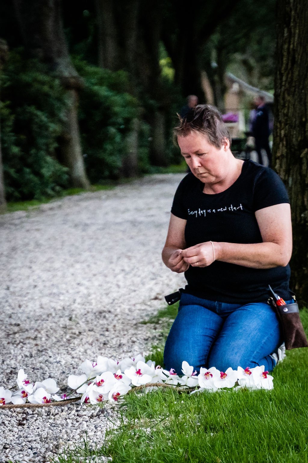 Een vrouw knielt op de grond naast een tak met witte orchideeën, met haar hoofd naar beneden en lijkt te bidden of te mediteren in een bosrijke omgeving.