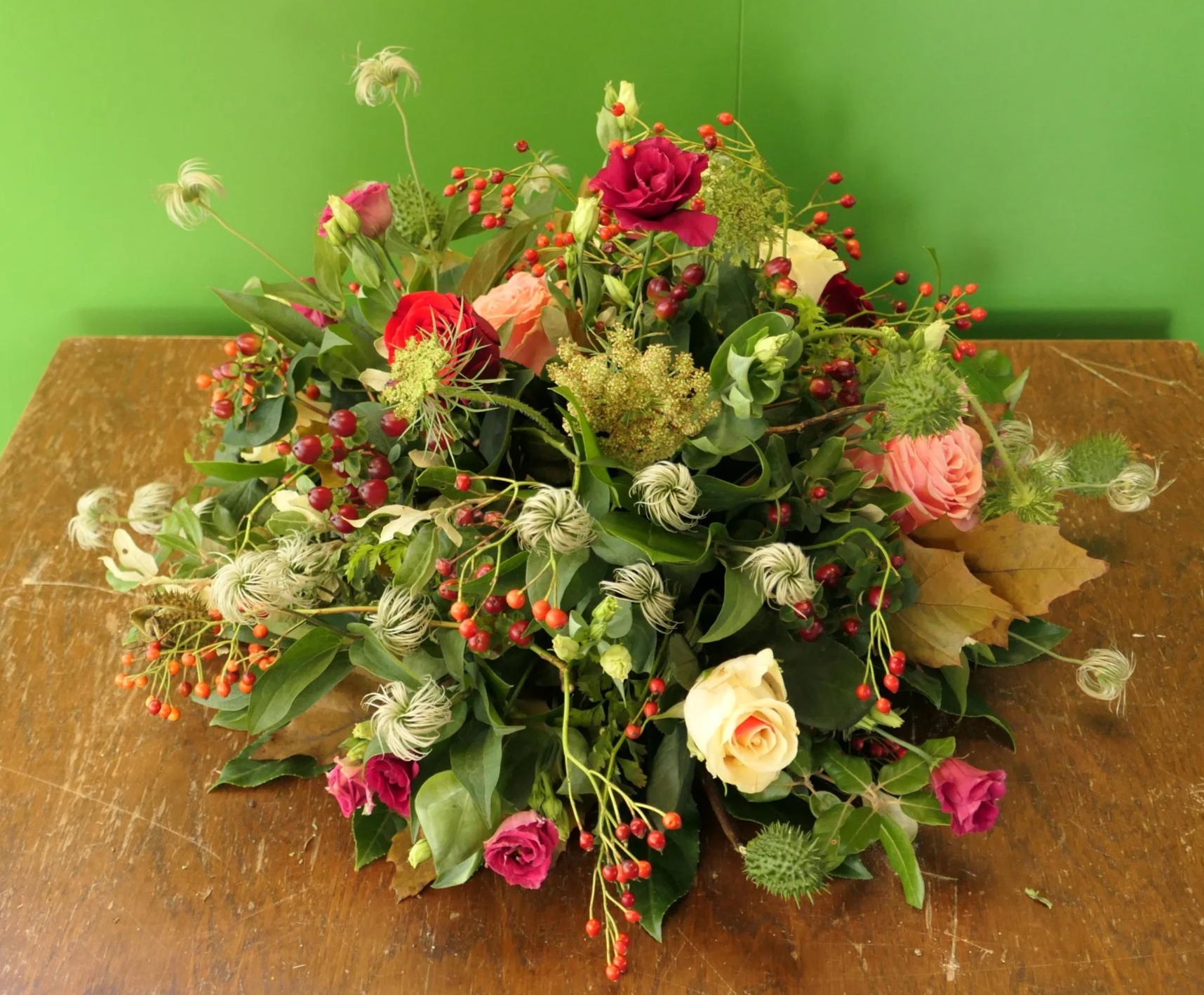 Een bloemenarrangement met rode, roze en witte bloemen, groene bladeren en rode bessen, geplaatst op een houten tafel.