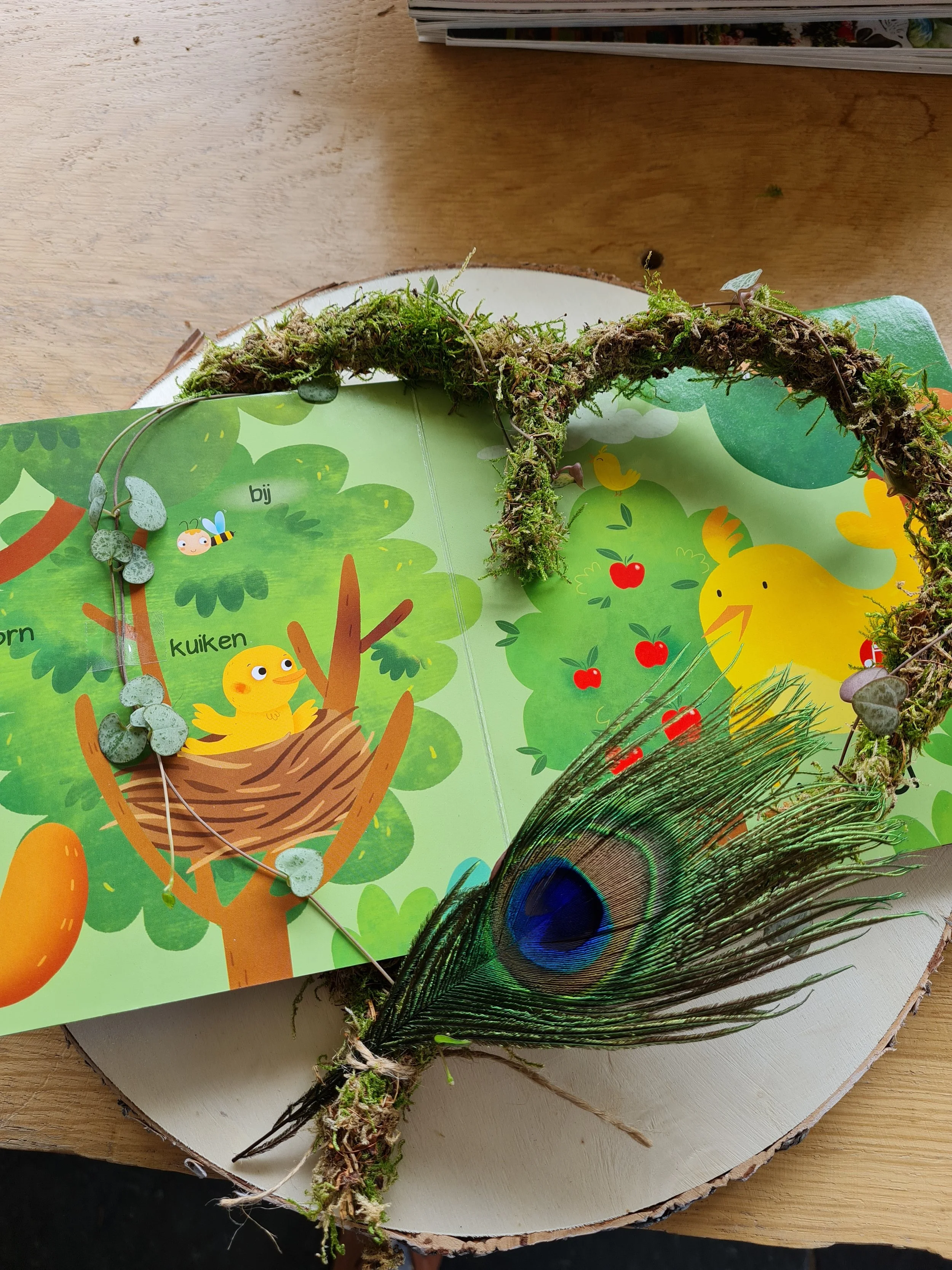 Een boek met een kleurrijke illustratie van een nest met een gele kuikentje, een boom en een bij, met een kip, en een pauwenveren naast het boek.