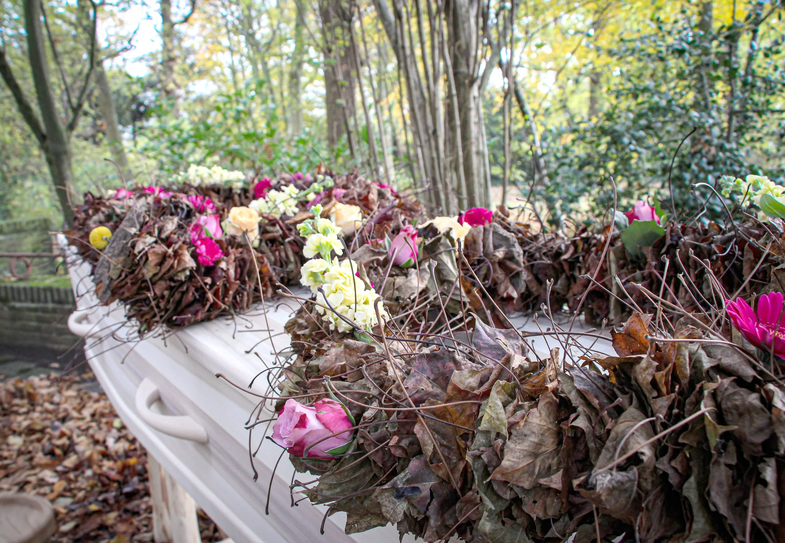 Een wit plastic rijsttafel met bladeren en bloemen, geplaatst op een bankje in een bosrijke omgeving met bomen en herfstkleuren.