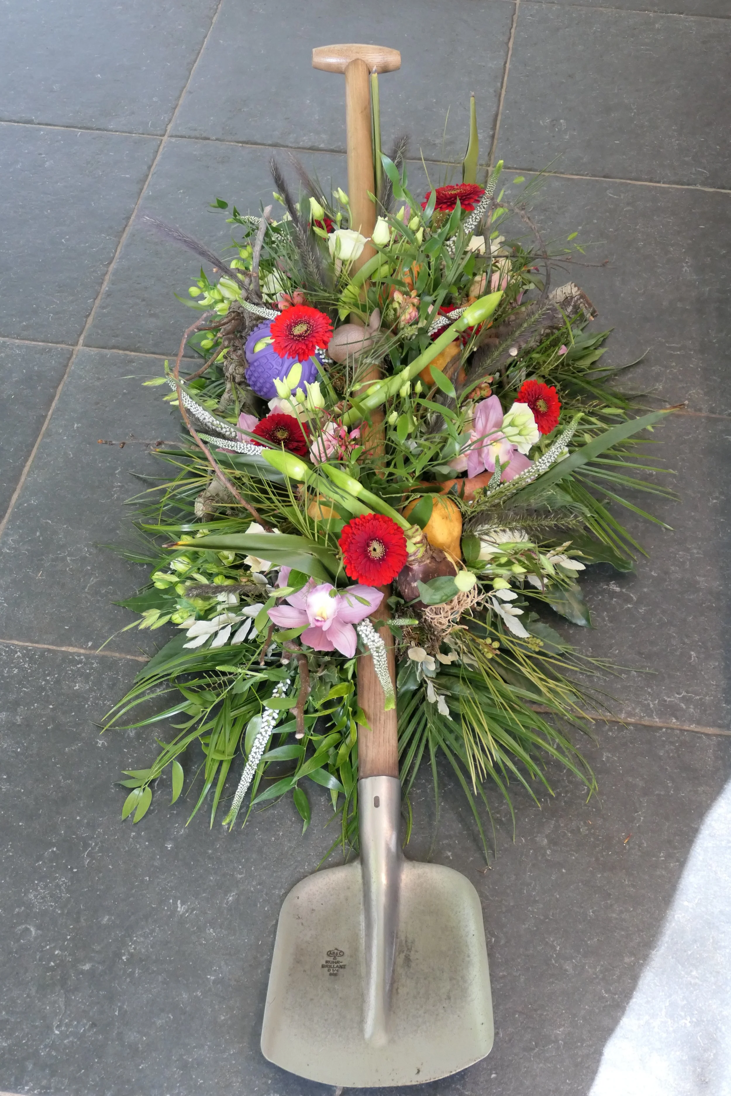 Een bezem met een bundel kleurrijke bloemen ligt op de vloer. De bloemen bevatten rode, witte, roze en paarse bloemen, vergezeld van groen blad en lange grassen.
