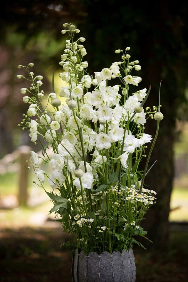 Een boeket witte bloemen in een grijze vaas, geplaatst in een tuin of bosrijke omgeving.