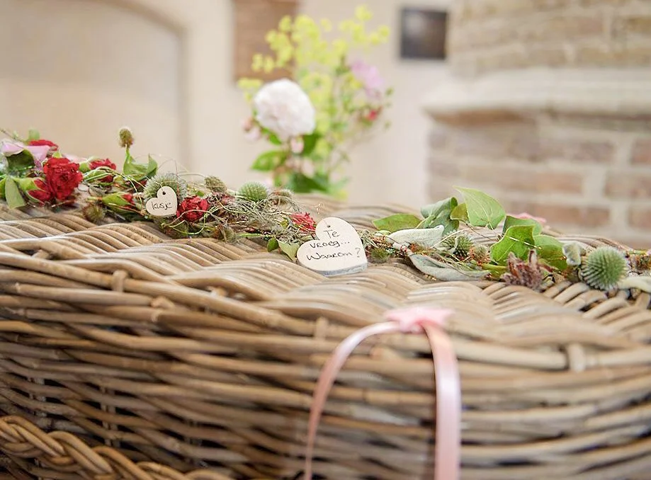 Een rieten mand met bloemen en kaarten, waarschijnlijk voor een bruiloft, met een bloementafel op de achtergrond.