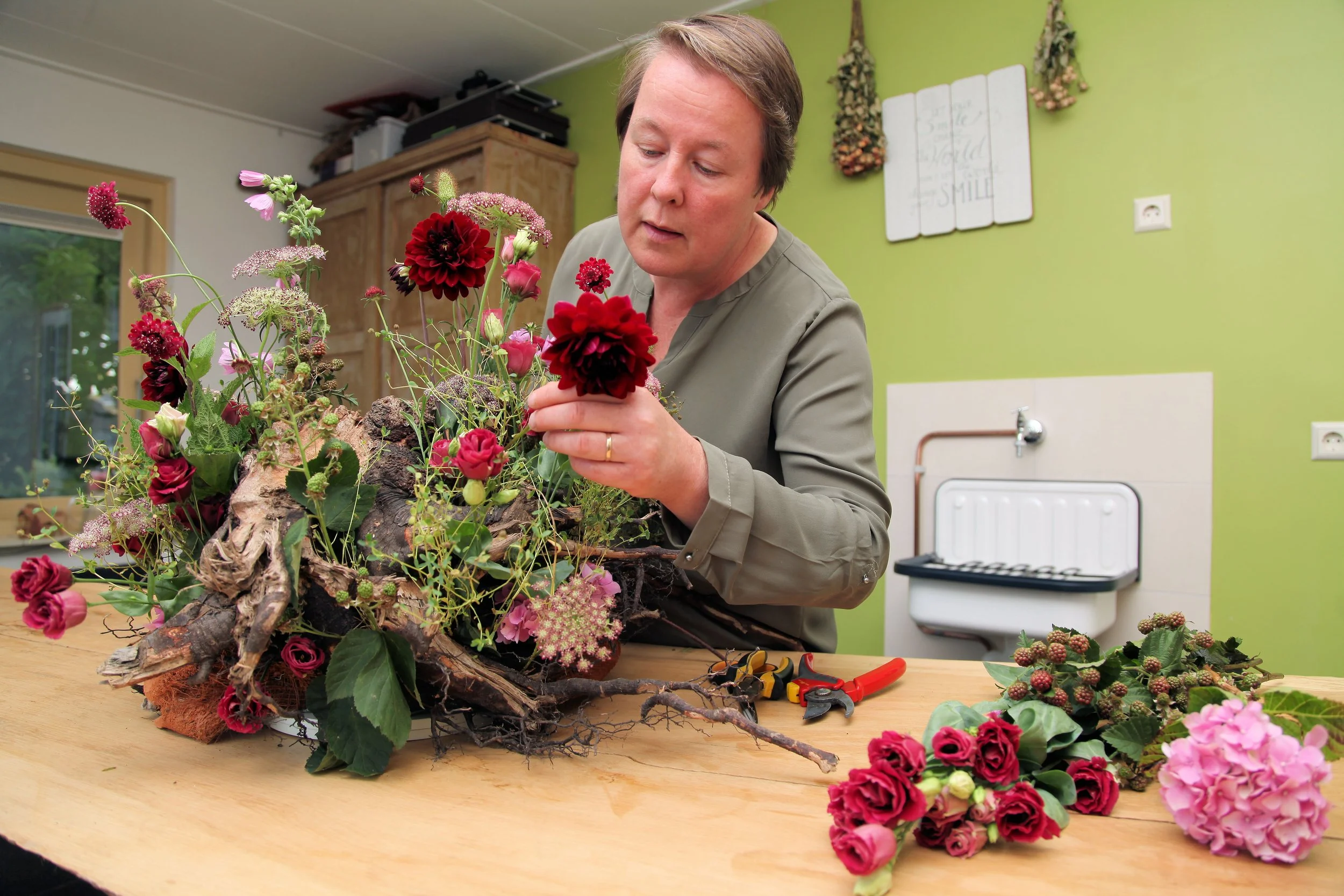 Man creëert een bloemstuk met kleurrijke bloemen, takken en planten op een houten tafel, met tuingereedschap op de voorkant.