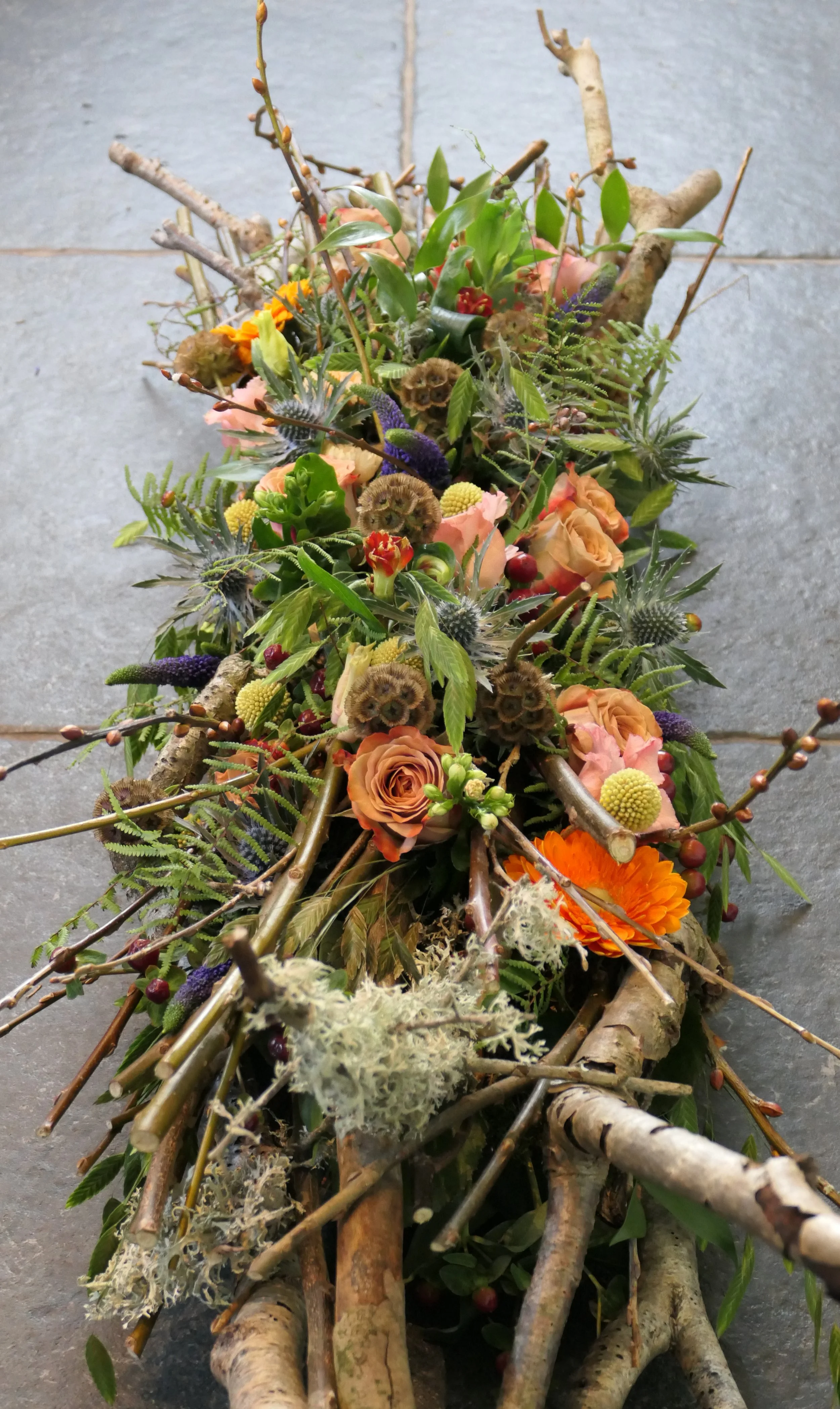 Kunstwerk bestaande uit taartspelden en natuurlijke materialen zoals takken, bladeren, bloemen en mos, vormgegeven als een bloemendecoratie op een vloer.