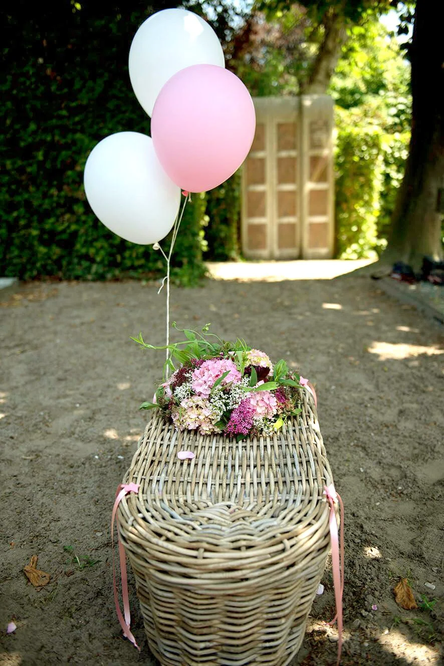 Een mand met bloemen en drie ballonnen, twee wit en één roze, op een zandachtige vloer in een tuin met groene planten en een houten hek in de achtergrond.