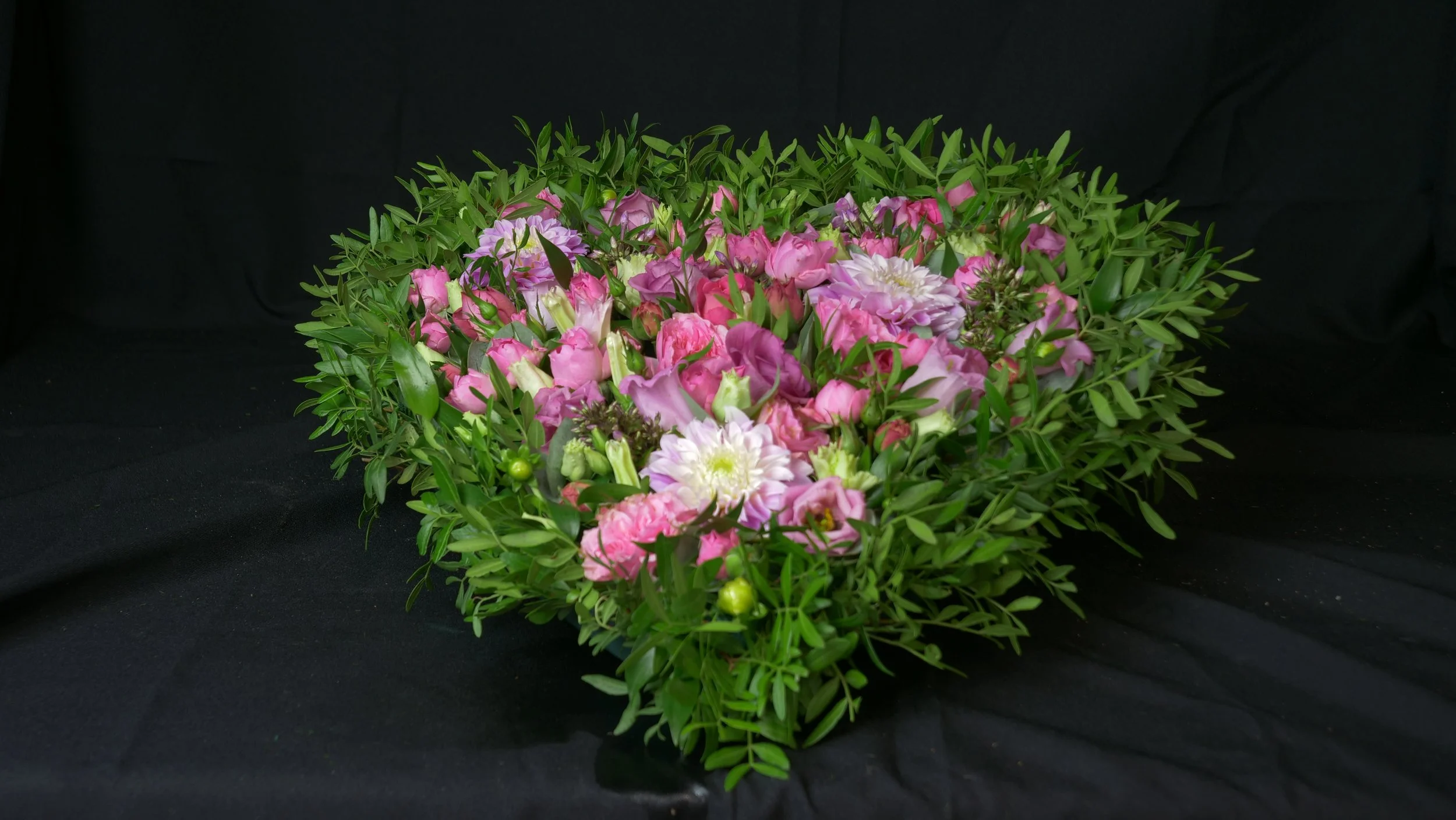 Een bloemstuk met diverse roze, paarse en witte bloemen omgeven door groene bladeren op een zwarte achtergrond.