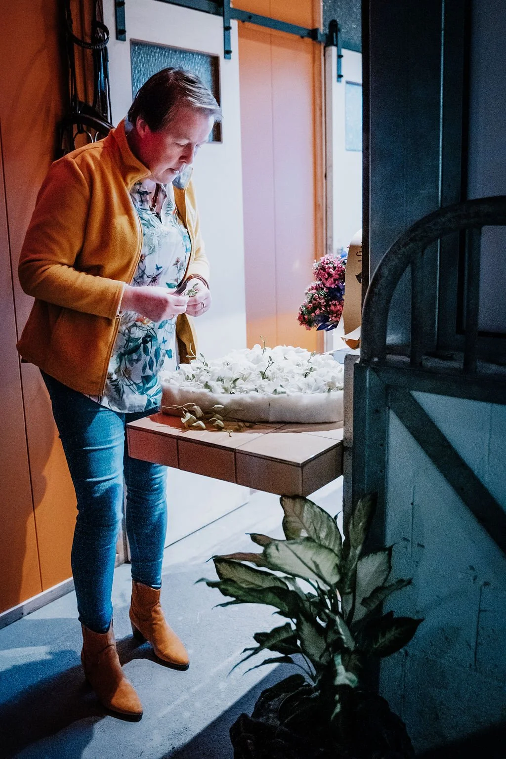 Vrouw in een geel-oranje jas en blauwe jeans plaatst witte bloemen in een bloemstuk.