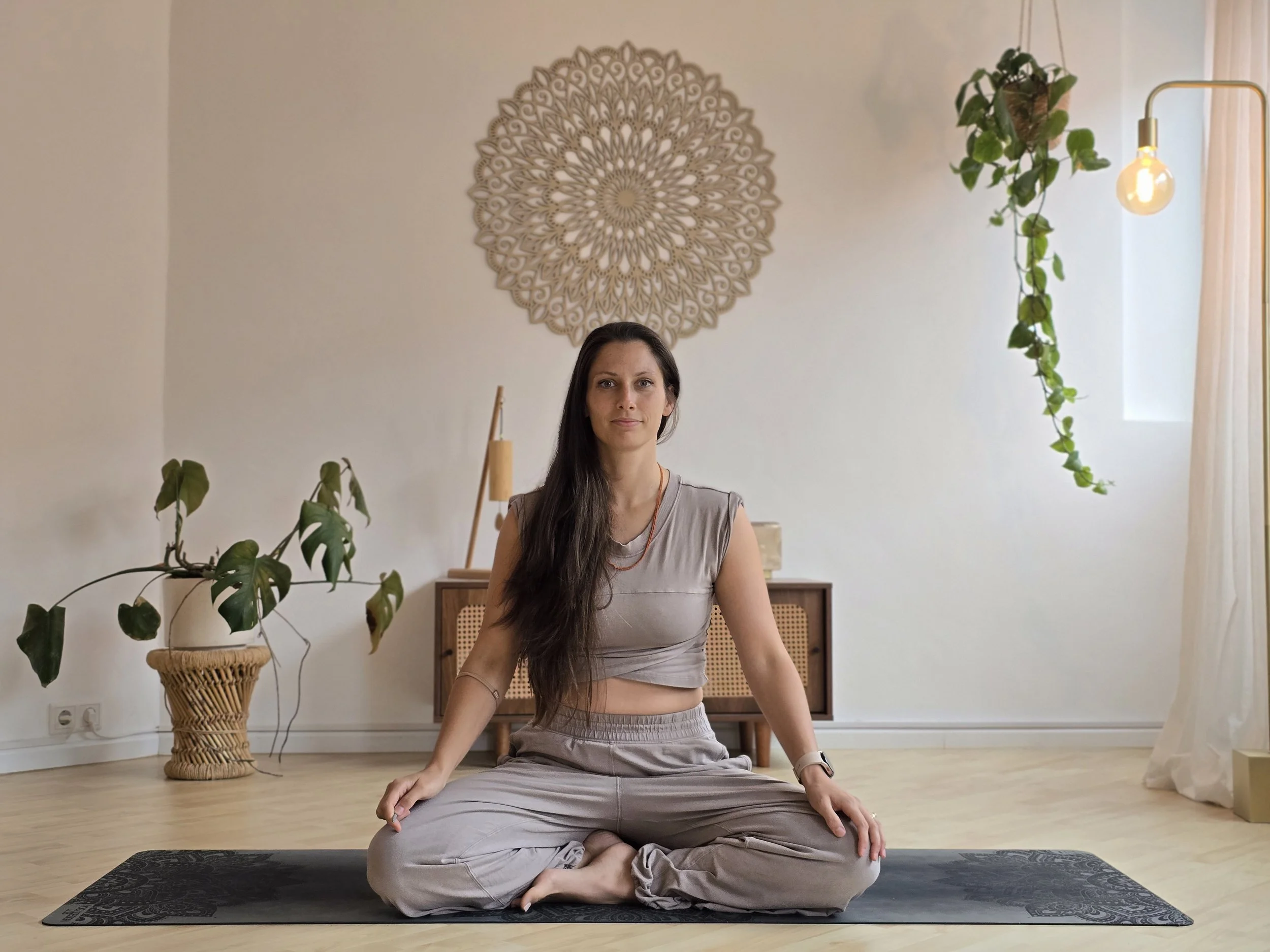 Eine Frau sitzt im Schneidersitz auf einer Yogamatte in einem hellen, modernen Raum mit Pflanzen, einem Wandbild und einem offenen Fenster mit Vorhängen.