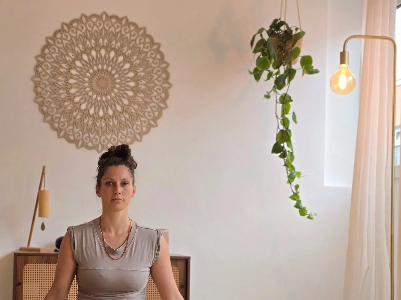 Eine Frau sitzt im Raum, im Hintergrund eine Wanddekoration, ein Holzregal, eine hängende Pflanze und eine Stehlampe mit Glühbirne, vor einem Fenster mit weißen Vorhängen.