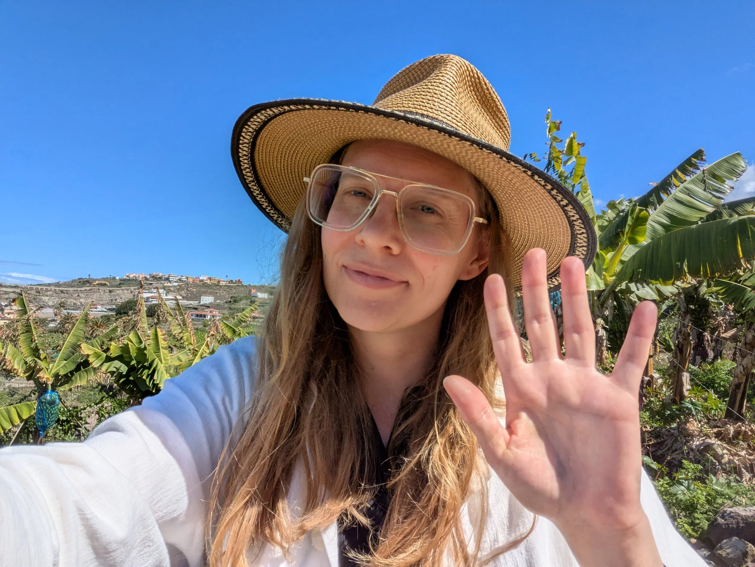 Young woman wearing a wide-brimmed straw hat and large glasses, smiling and waving outdoors against a clear blue sky and lush green vegetation.
