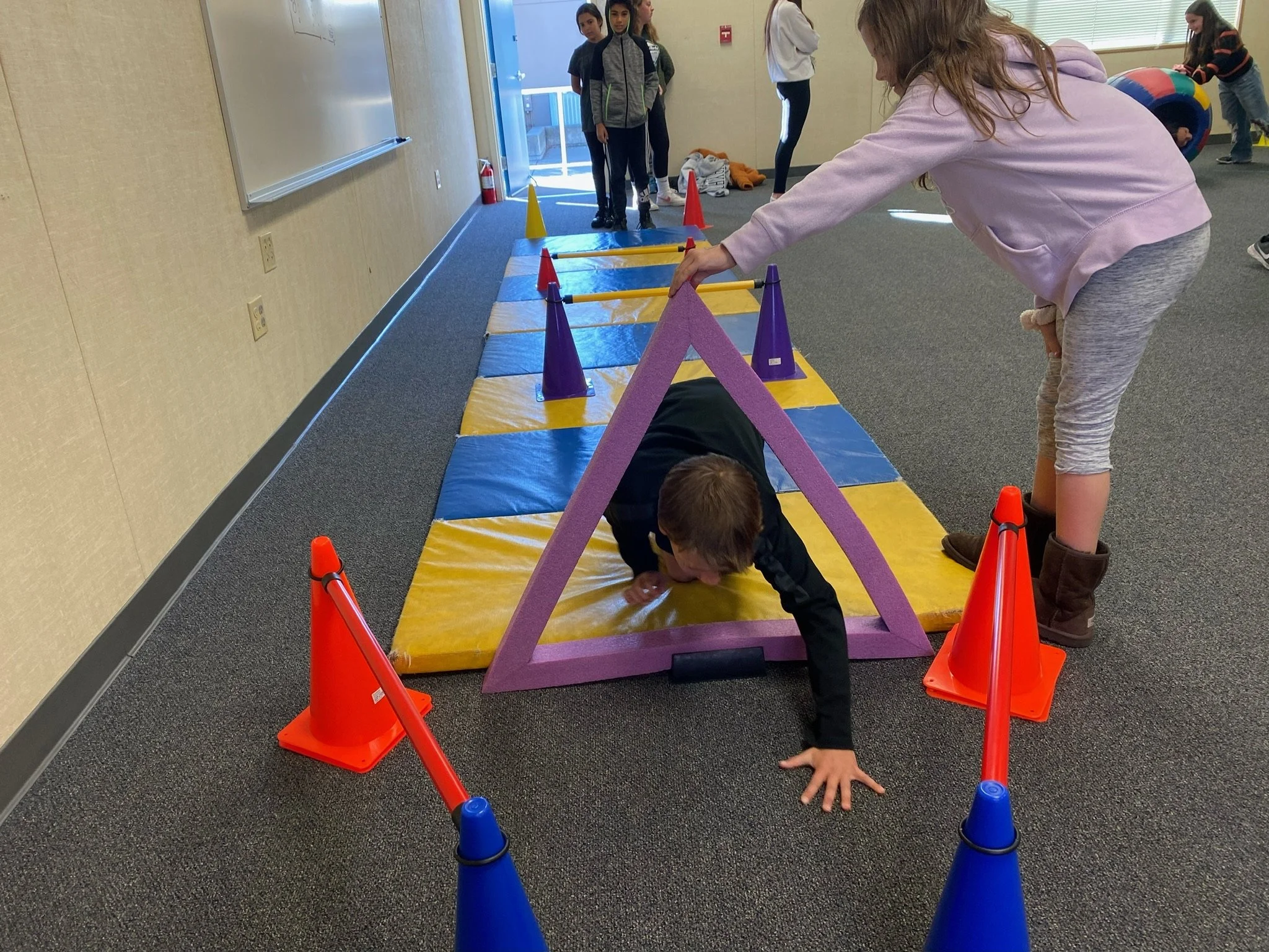 Children participating in a school indoor activity, crawling through a purple foam triangle obstacle, surrounded by orange and blue cones, on a blue and yellow mat, with other children in the background.
