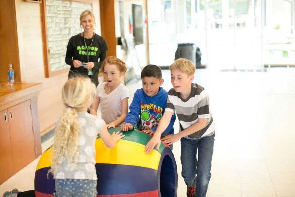 A group of children pushing a colorful cylindrical object while a woman watches and smiles in the background.