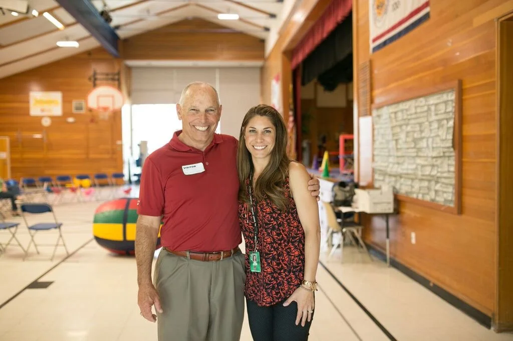 A man and a woman standing together inside a gymnasium or indoor sports facility, smiling at the camera. The man is wearing a red polo shirt and khaki pants, and the woman is in a sleeveless patterned top and dark jeans.