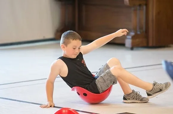 A young boy sitting inside a red plastic bowl, balancing with one arm on the floor and the other raised, on a light-colored gym floor.