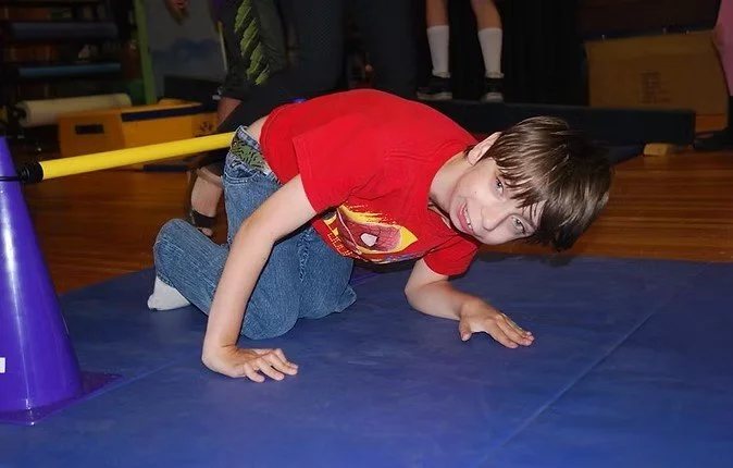 A young boy with brown hair in a red t-shirt and jeans on a blue mat, crawling on all fours with his tongue out, in an indoor play area.