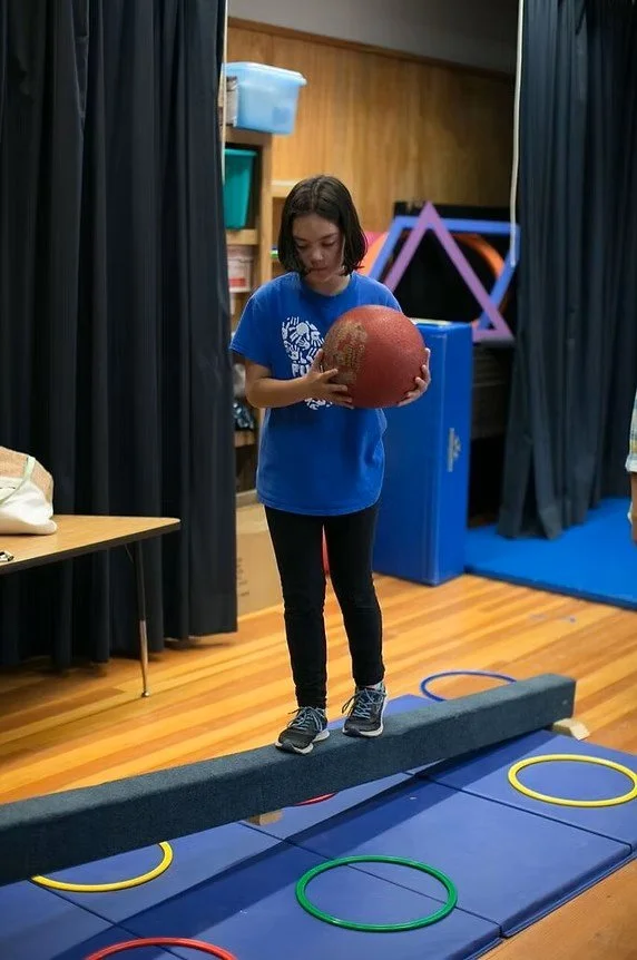 A girl in a blue shirt and black pants balances on a gymnastics beam while holding a red ball. She is in a room with wooden floors and black curtains, and there are colorful hoops on the floor in front of her.
