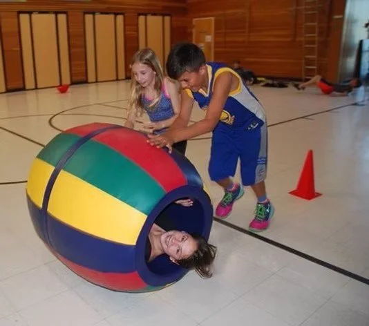 Two children are playing with a large, colorful inflatable ball in a gymnasium, with one child inside the ball and the other two pushing it.