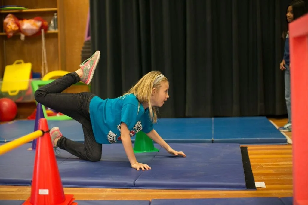 Young girl in a blue T-shirt and black pants performing an obstacle course on gym mats, balancing on her hands and knees with one leg lifted behind her, in a gym with colorful equipment and a black curtain in the background.