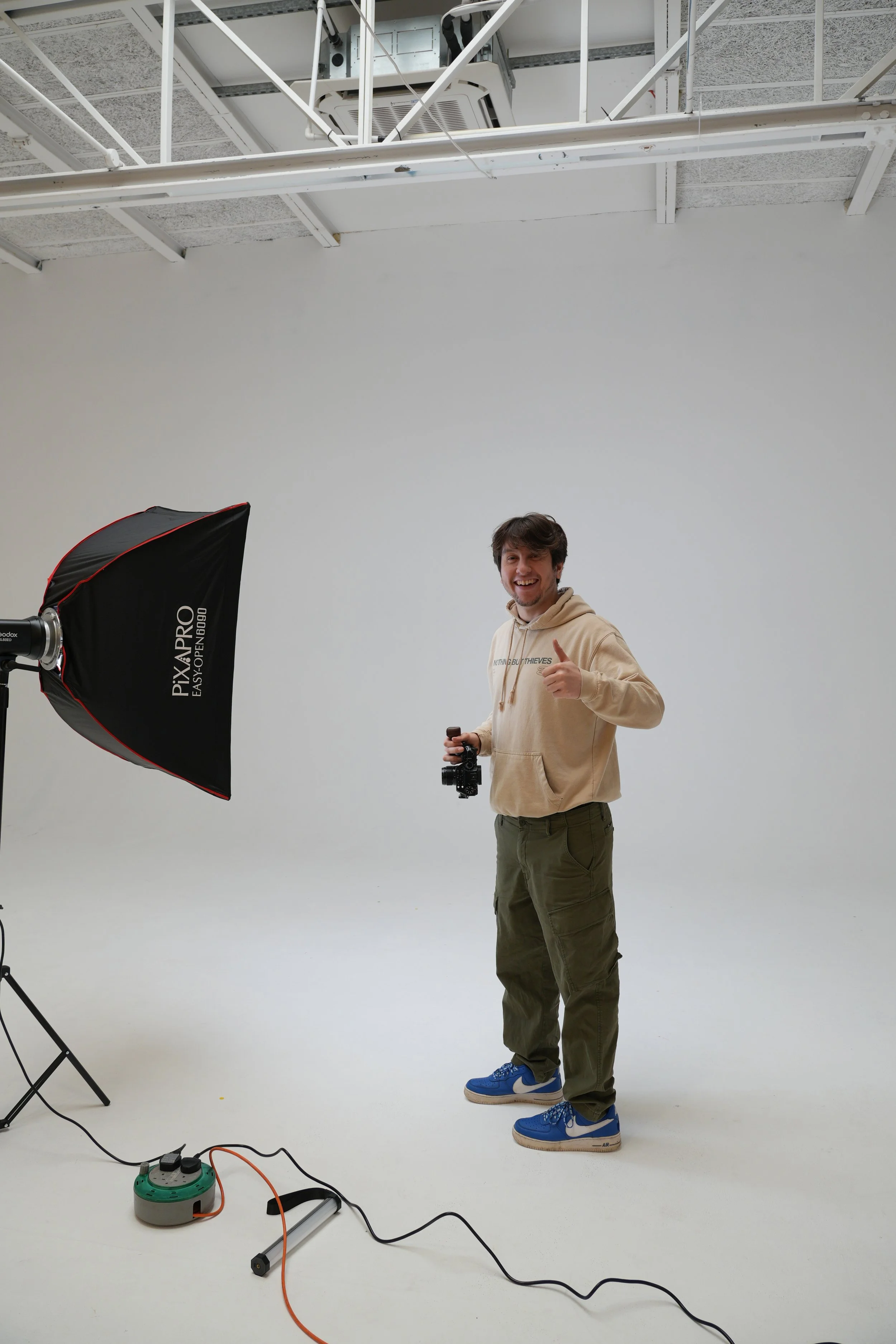 A young man in a beige hoodie and green cargo pants standing in a photography studio, holding a camera and smiling while giving a thumbs-up. There is a large studio light with a softbox and some electrical equipment on the floor. The background is plain white.