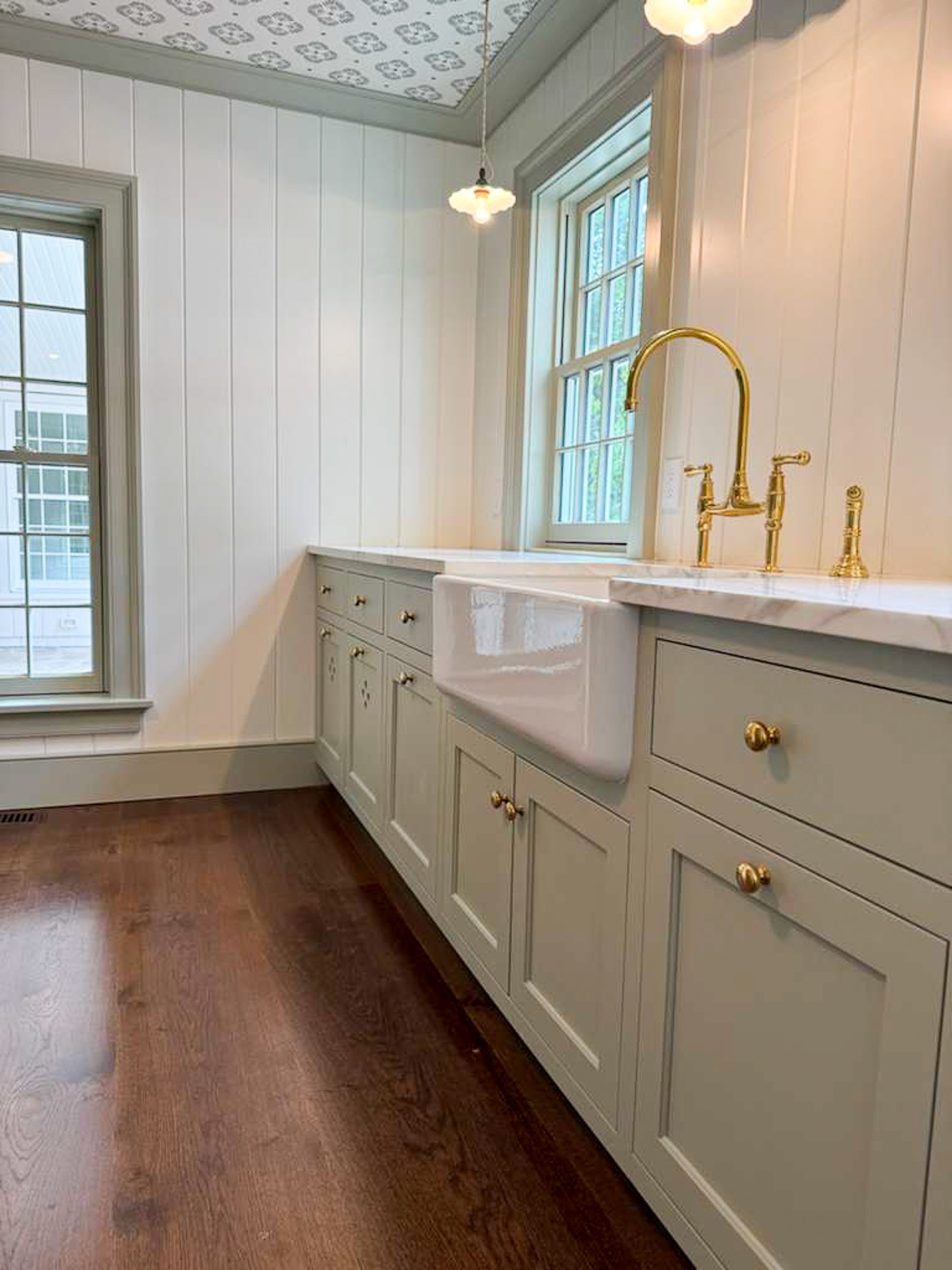 A kitchen with white wood-paneled walls, large windows, and a vintage farmhouse sink with gold fixtures. There are hardwood floors and a paper ceiling with floral patterns, along with small pendant lights hanging from the ceiling.