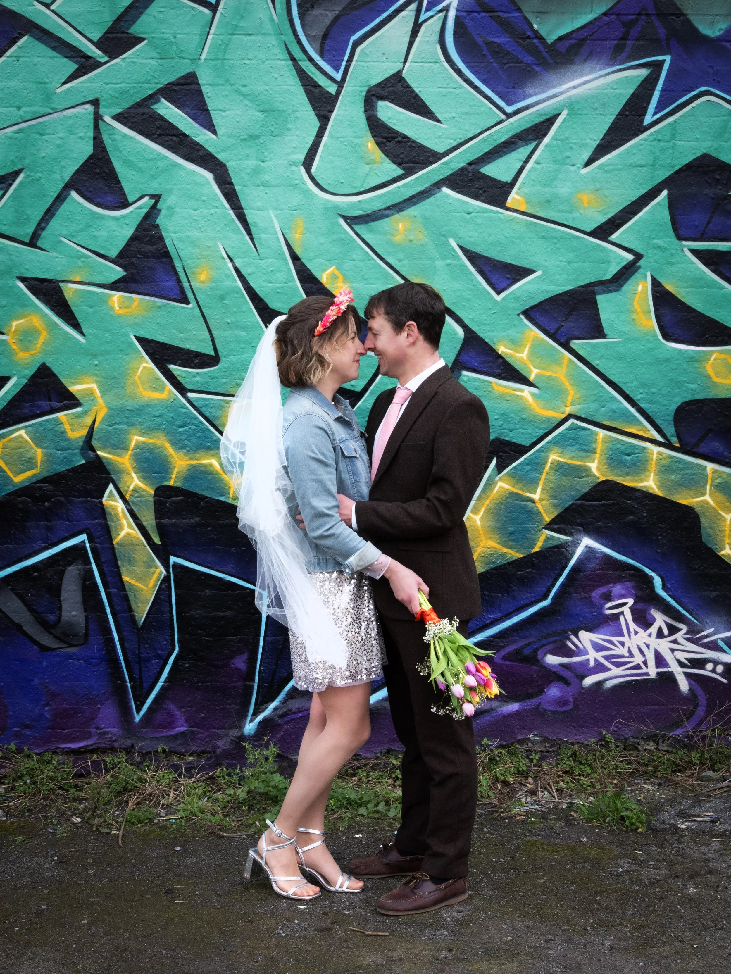 A wedding couple standing close together with their foreheads touching, holding a bouquet of tulips, in front of a colorful graffiti wall. Bristol Dean Lane Skatepark